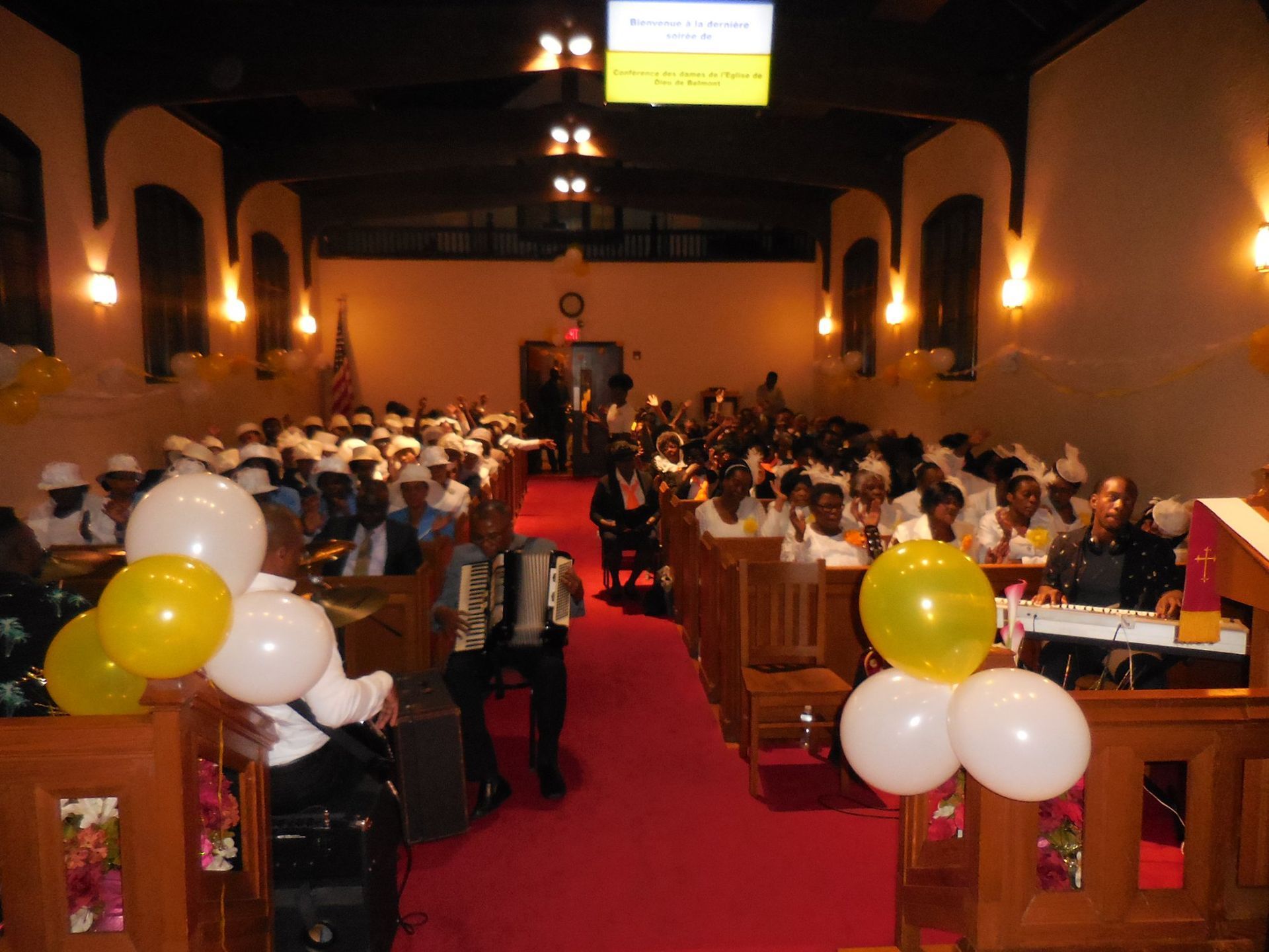 A church congregation sits in wooden pews facing a red aisle, decorated with white and yellow balloons.