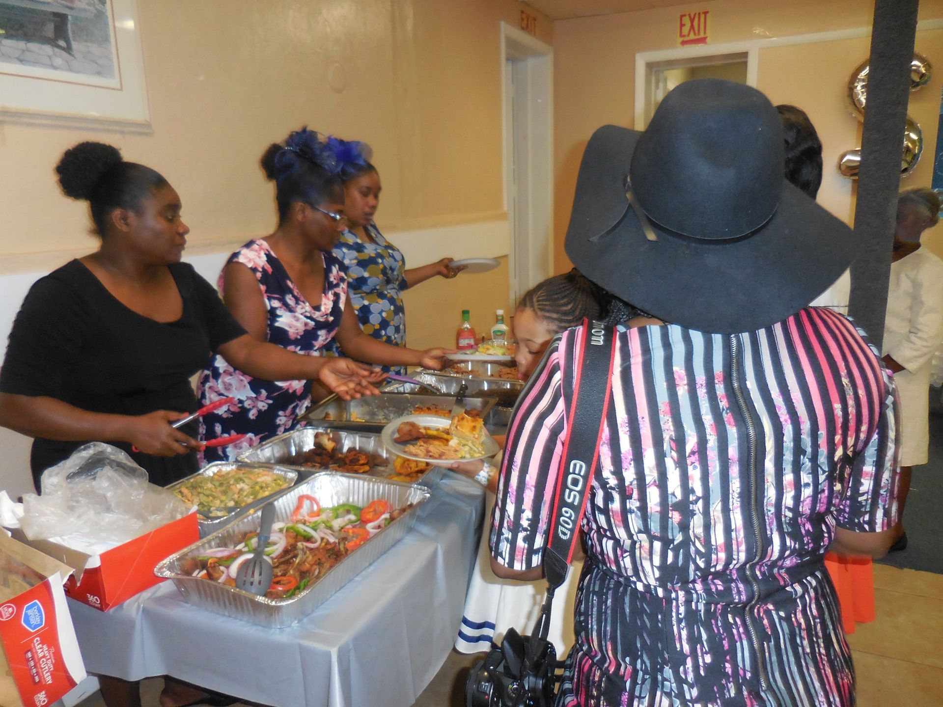 People in a serving line take food from trays laid out on a table at an indoor event.