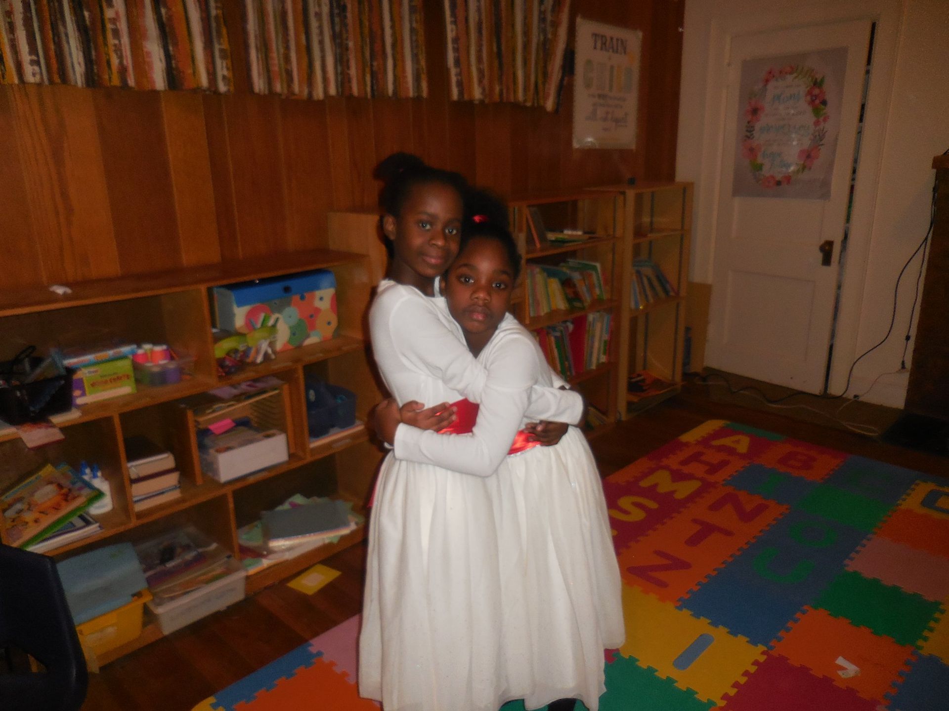 Two children in white dresses embrace in a room with wooden walls, shelving units, and a colorful alphabet floor mat.