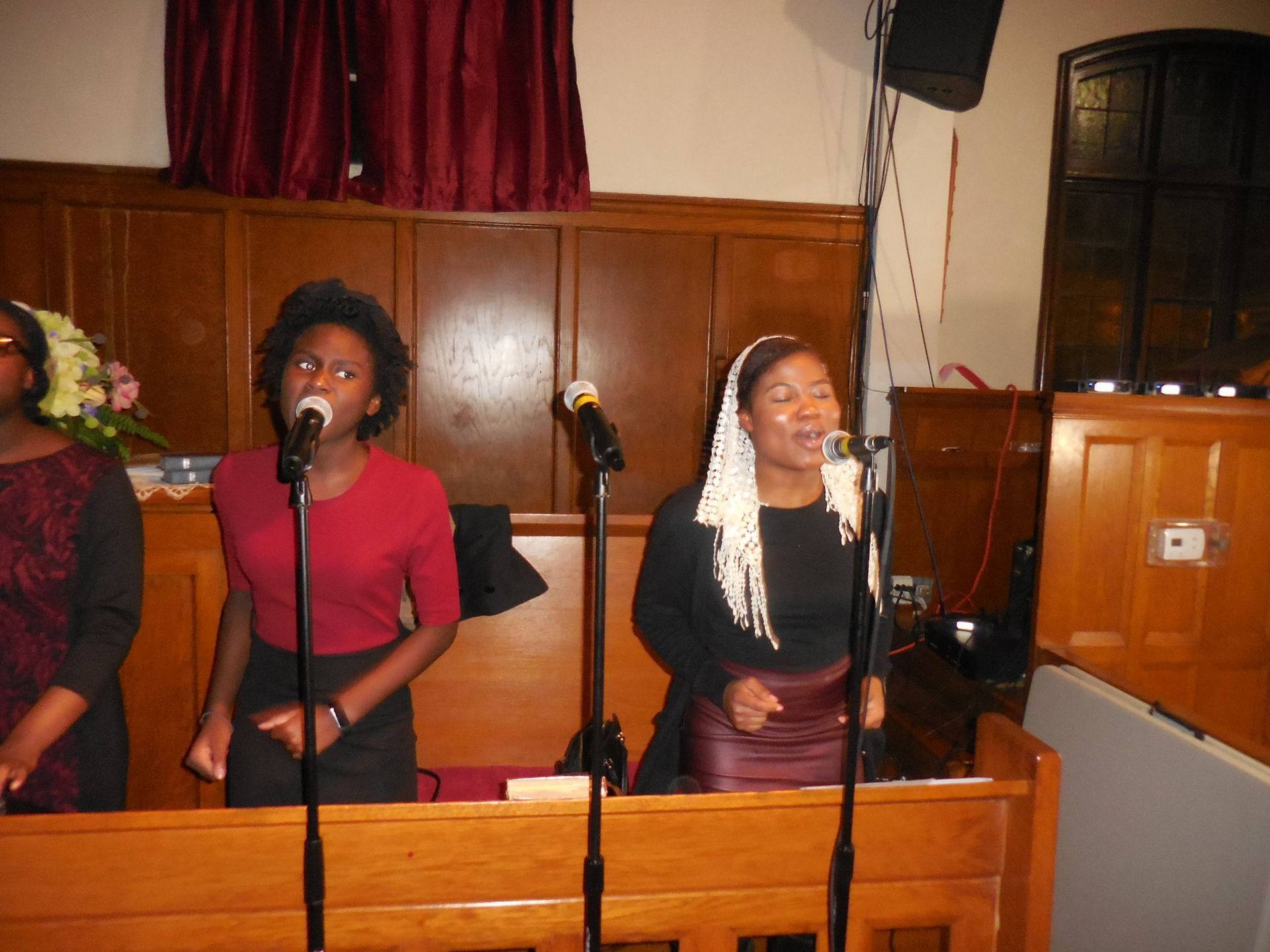 Three people singing into microphones in front of wood-paneled walls in a church setting.