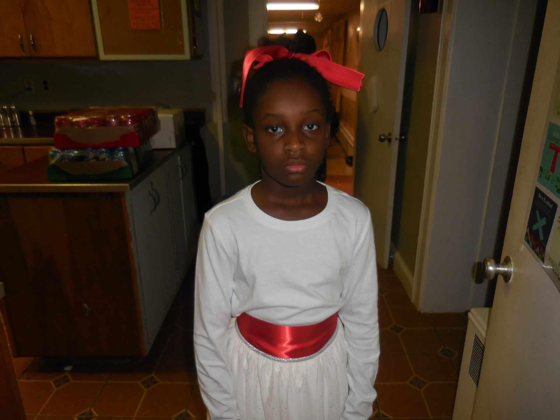 A person wears a white long-sleeved shirt with a red sash and hair bow, standing in a room with wood cabinets.