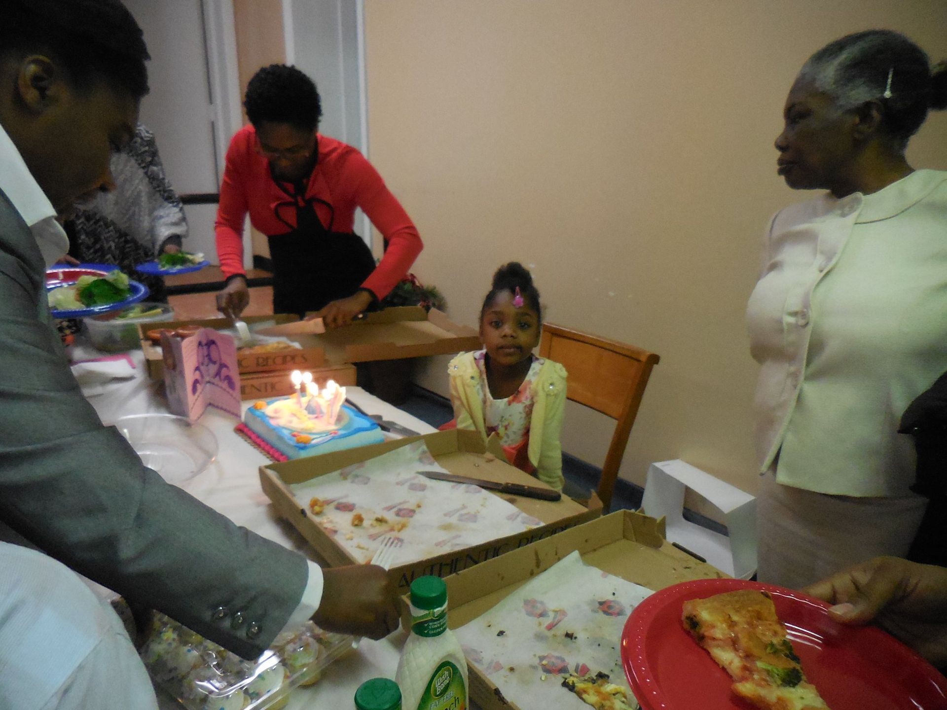 A group of people gather around a table with a small birthday cake and boxes of pizza for a celebration.