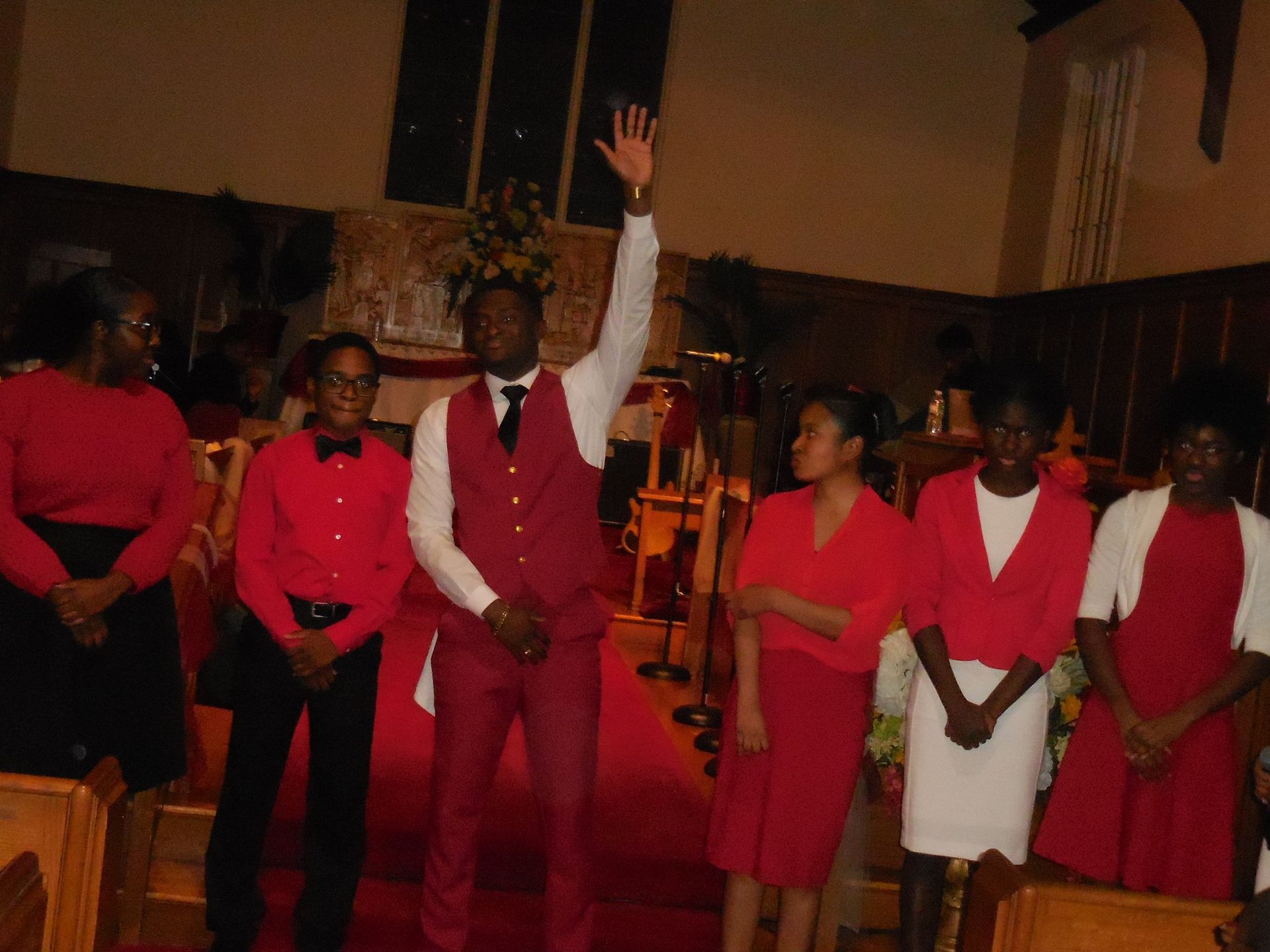 A group in red and white formal wear stands in a church; one person raises a hand as if leading or speaking.