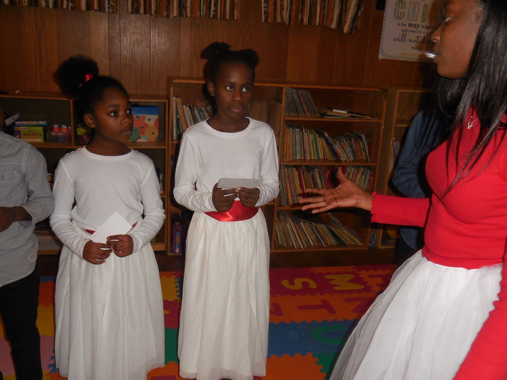 Two children in white dresses and a person in a red top stand in a room with wooden bookshelves and a colorful play mat.