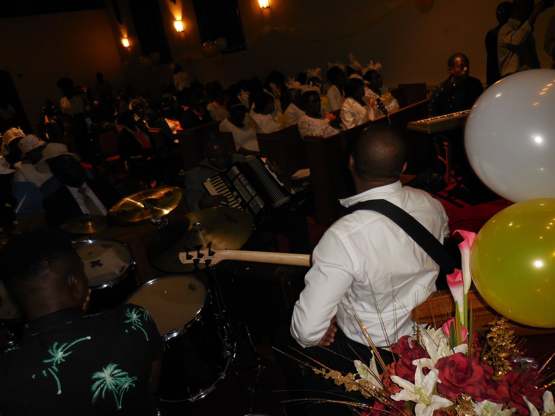 A band plays on stage at a dimly lit event, featuring a drummer and a guitarist beside a floral arrangement and balloons.