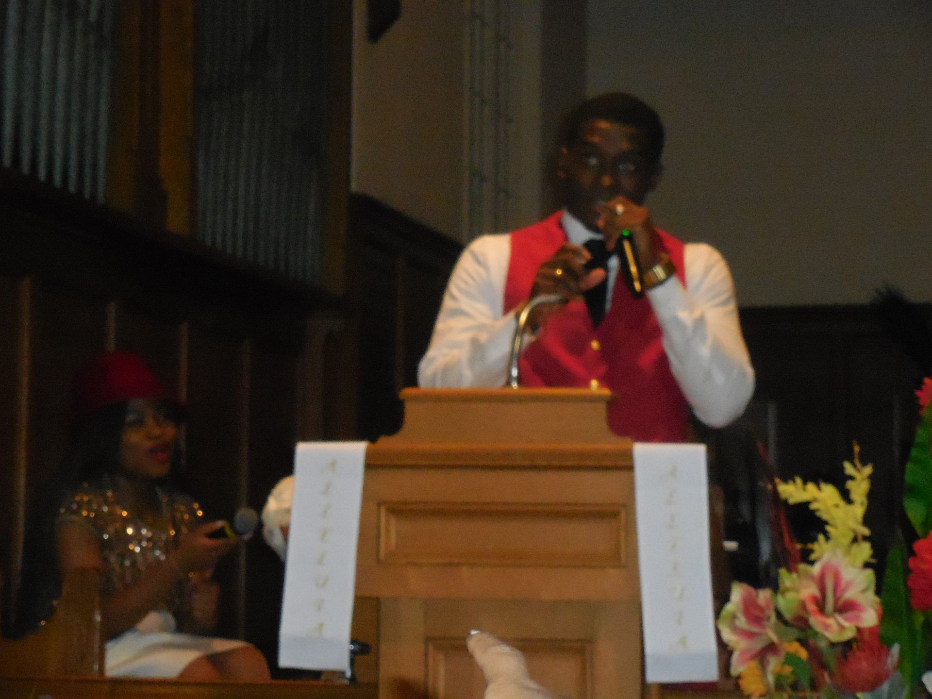 A speaker in a red vest and white shirt stands at a wooden pulpit, holding a microphone, with a person seated nearby.
