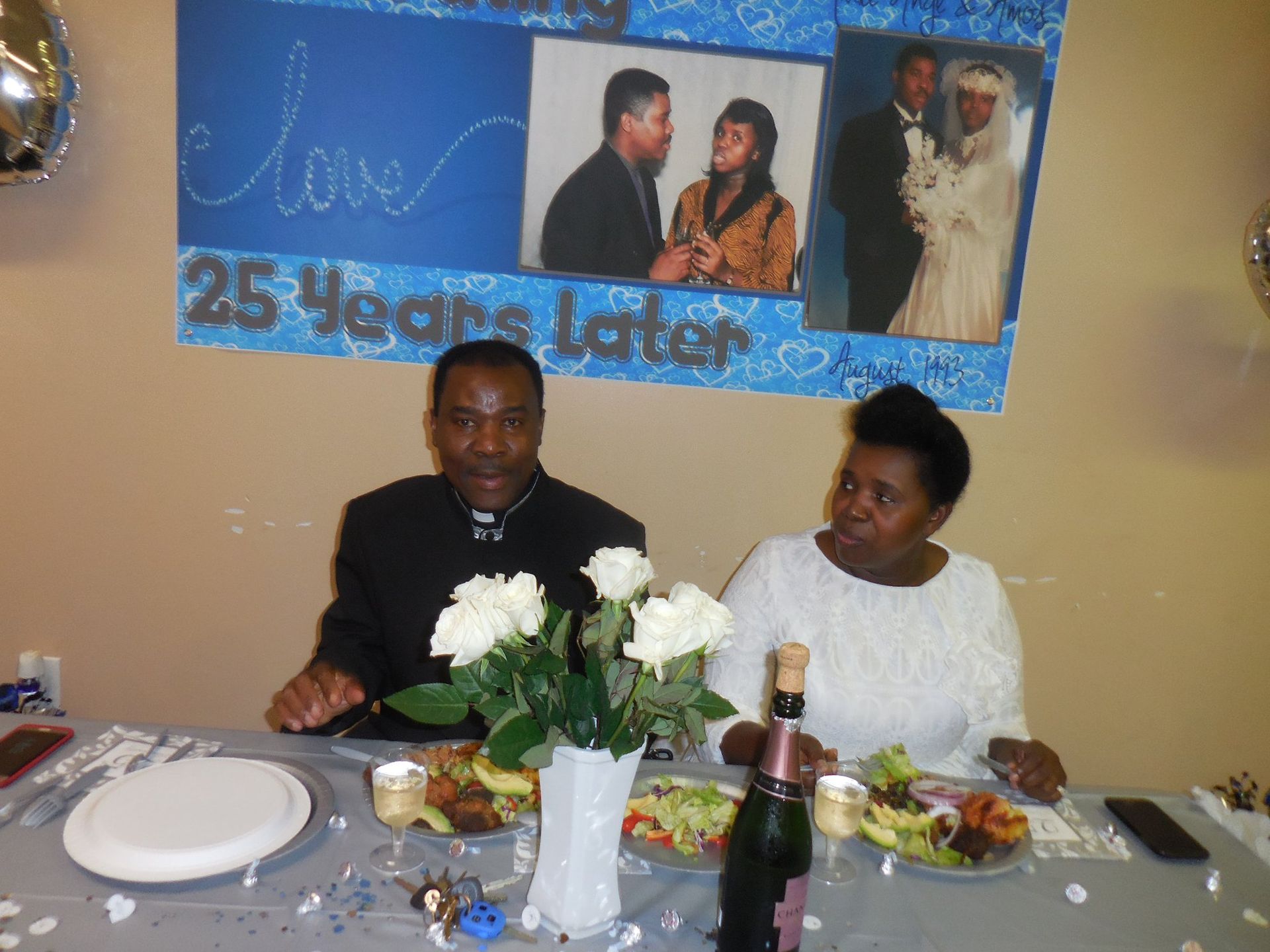 A couple sitting at a table decorated for their 25th anniversary, with photos of them from their wedding day displayed.