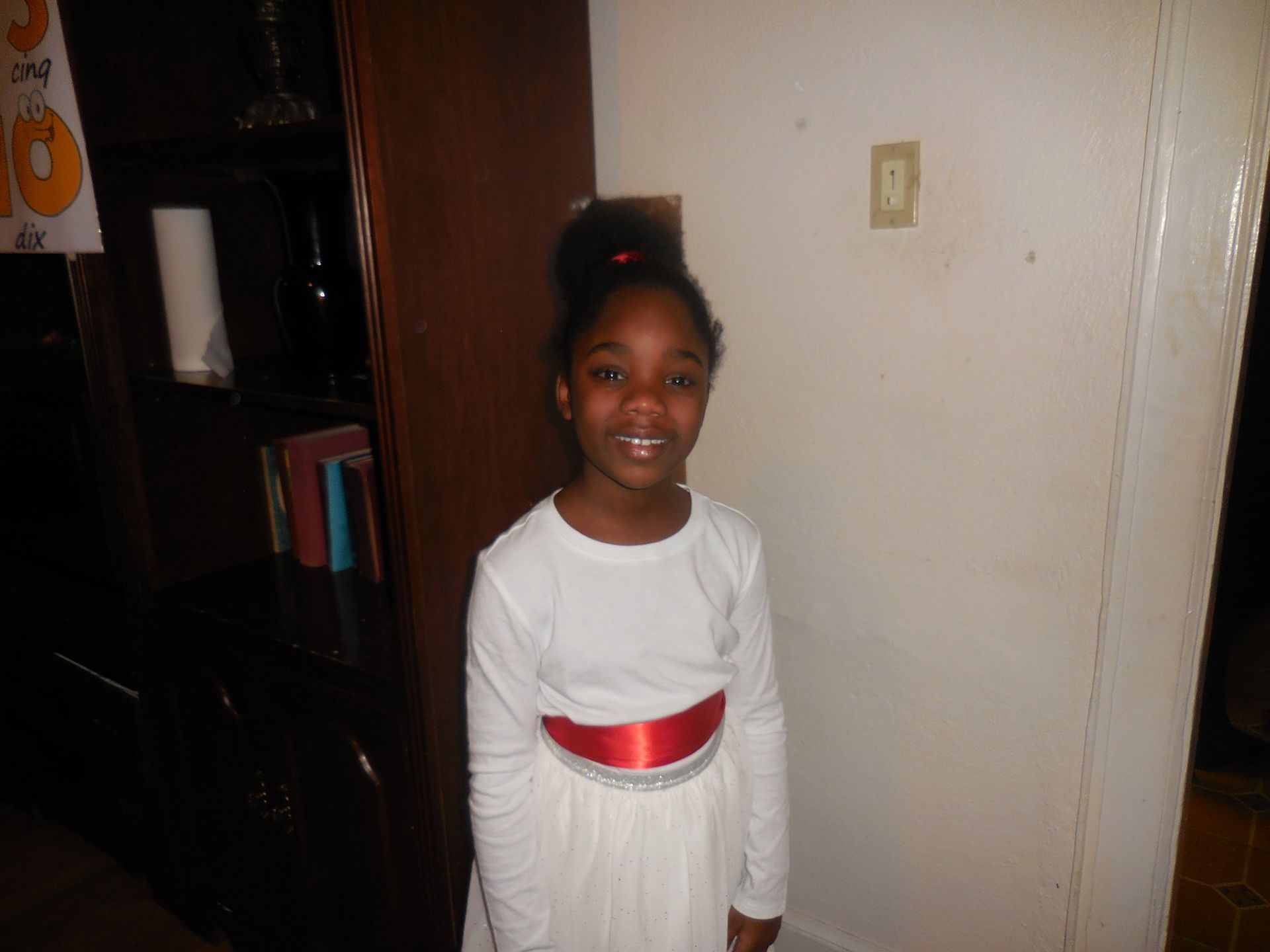 A smiling child wearing a white long-sleeved shirt with a red sash stands near a dark wooden shelf and a plain wall.