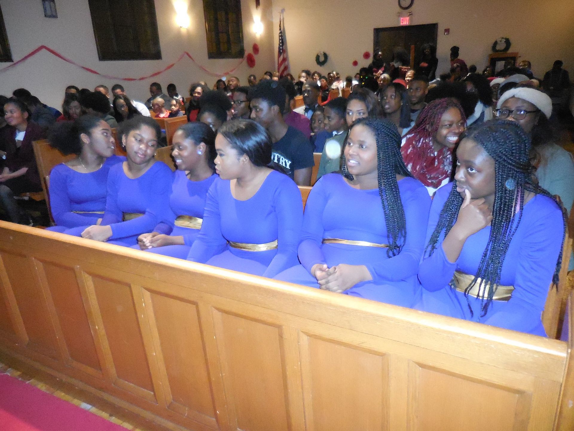 A row of people in matching blue dresses with gold belts sit in a church pew, with a congregation in the background.