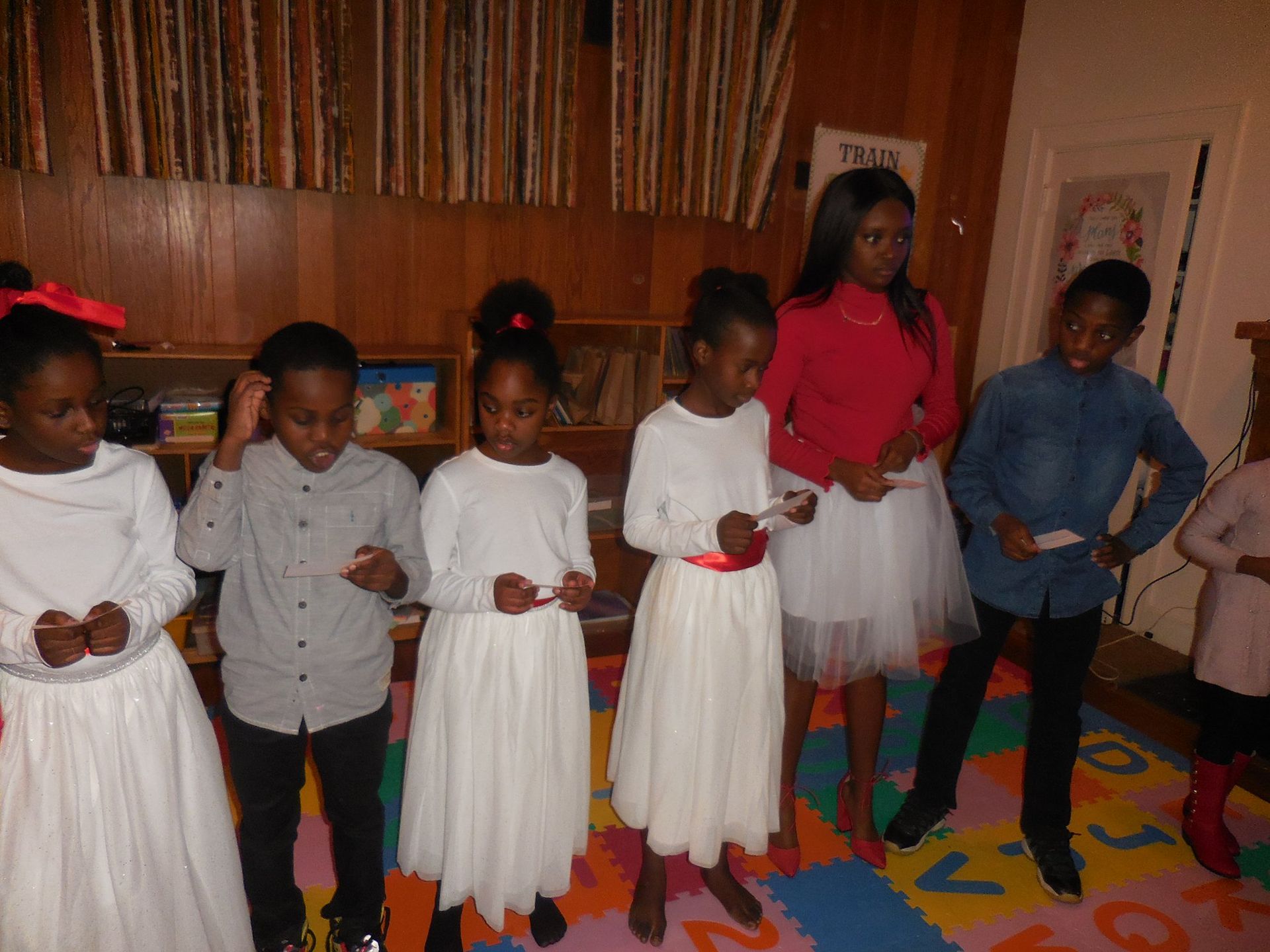 A group of people standing in a line indoors on a lettered rug, some holding papers as if preparing for a performance.