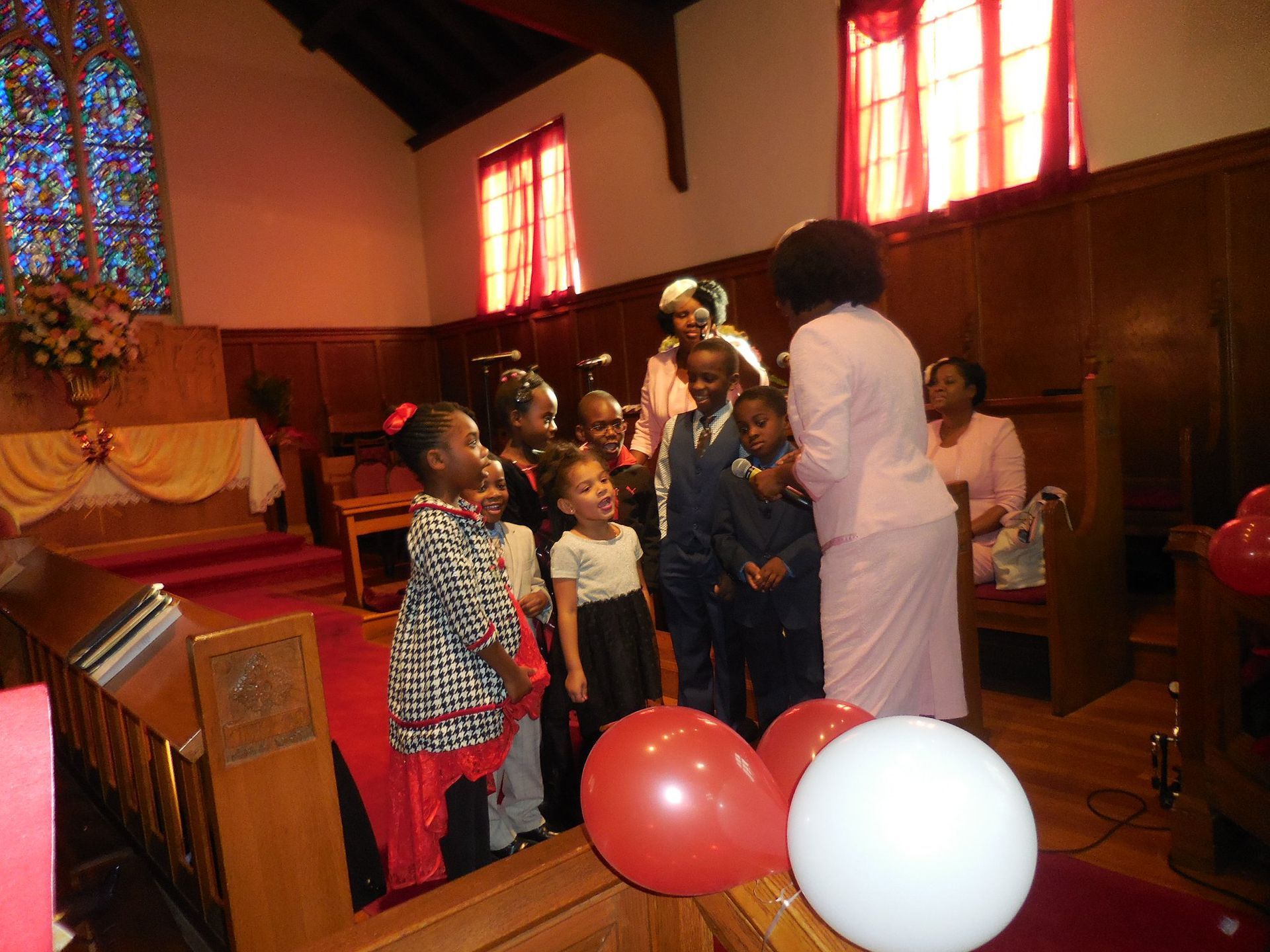 A group of children and adults standing in a church with red and white balloons in the foreground.