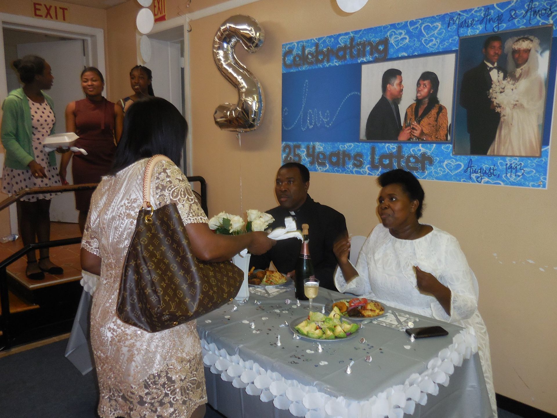 A couple sits at a table decorated for a 25th anniversary celebration, with guests nearby in a room with a wall banner.