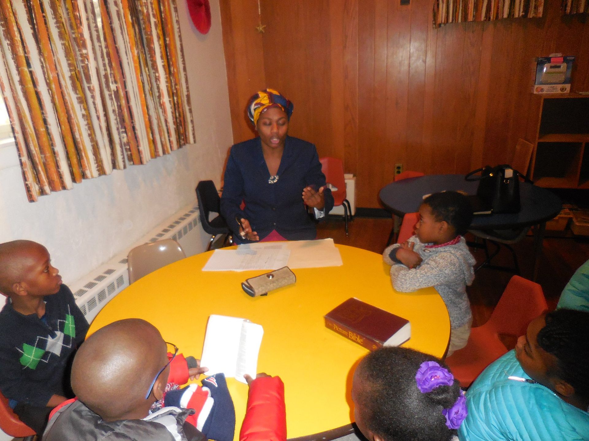 A teacher sits at a round yellow table, engaging with four children in a classroom with wood-paneled walls.