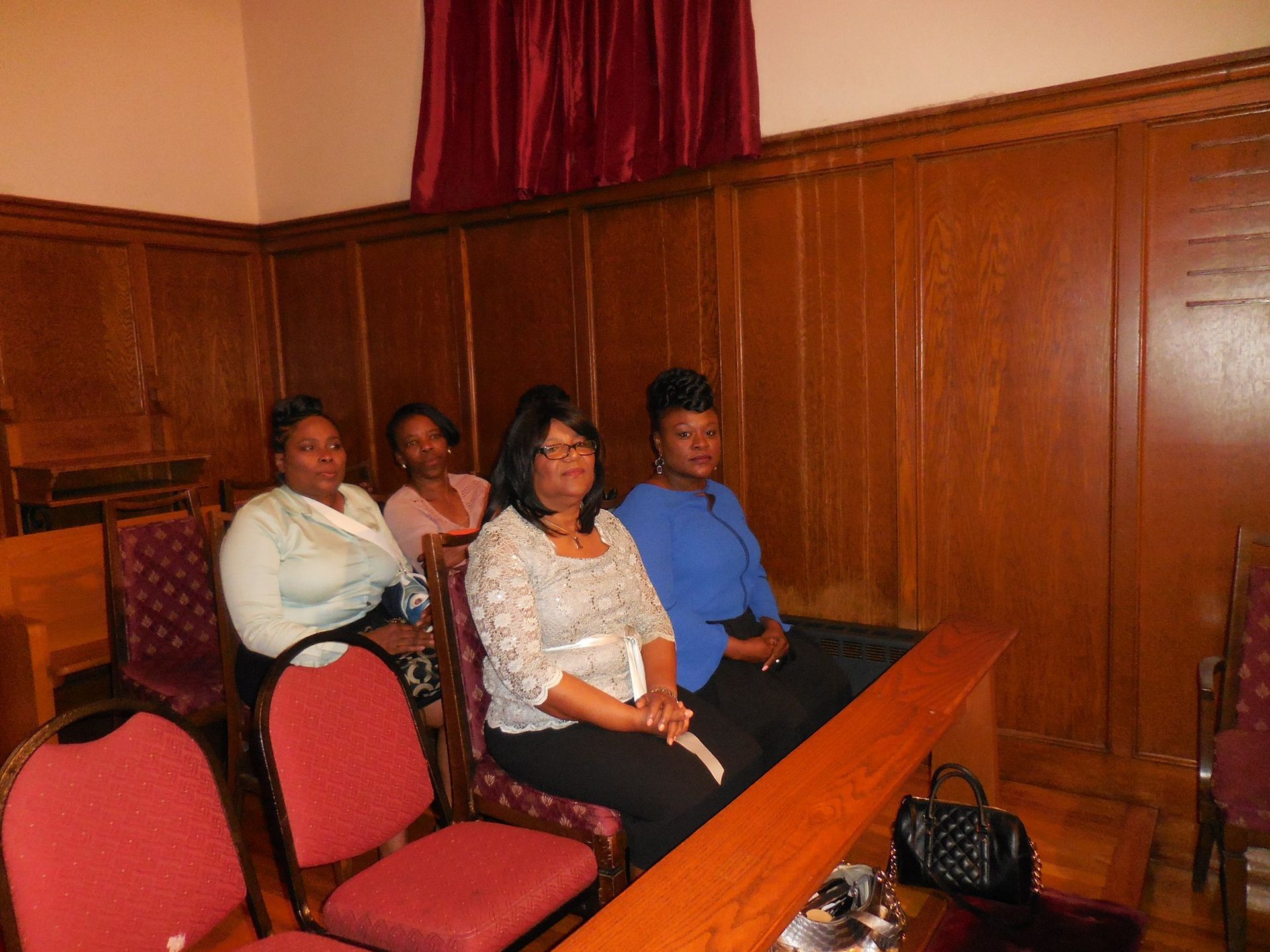 Four people sit in wooden chairs in a wood-paneled room with red curtains.