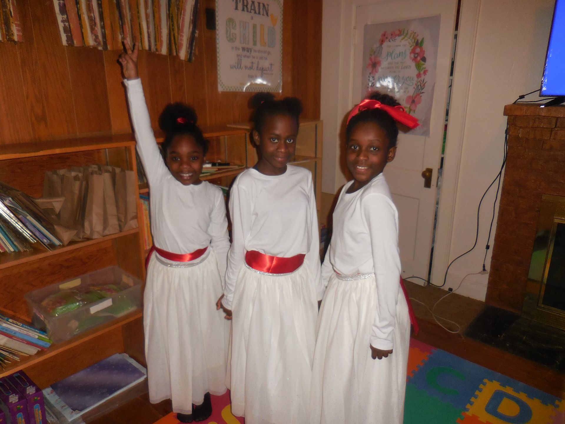 Three children in matching white long-sleeve shirts and long white skirts with red sashes standing indoors.