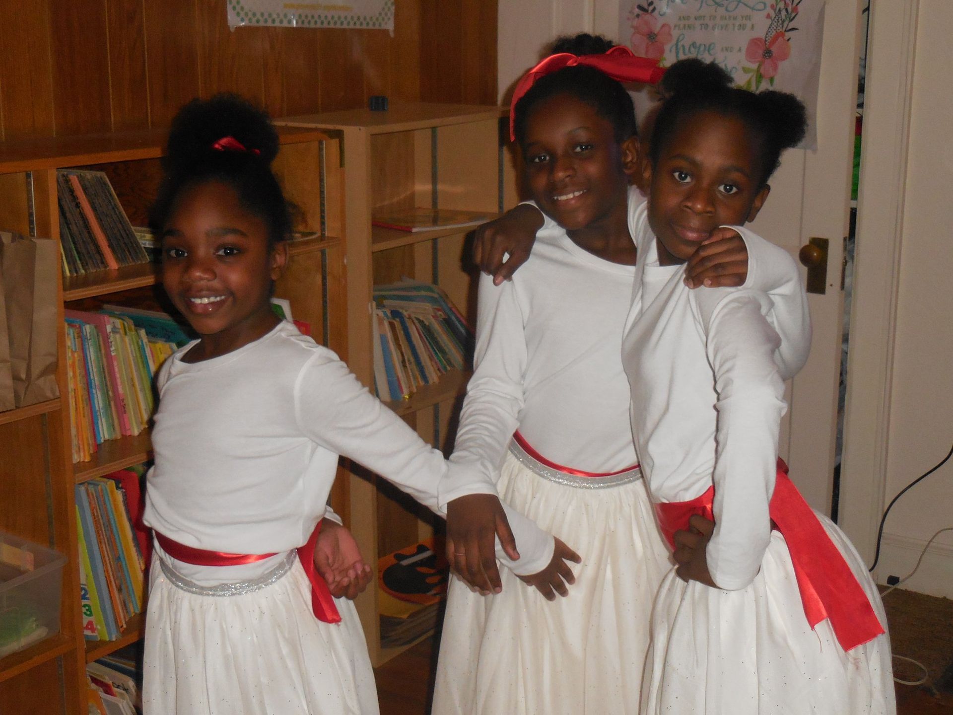 Three smiling children in white long-sleeved tops and white skirts with red sashes standing together in a room.