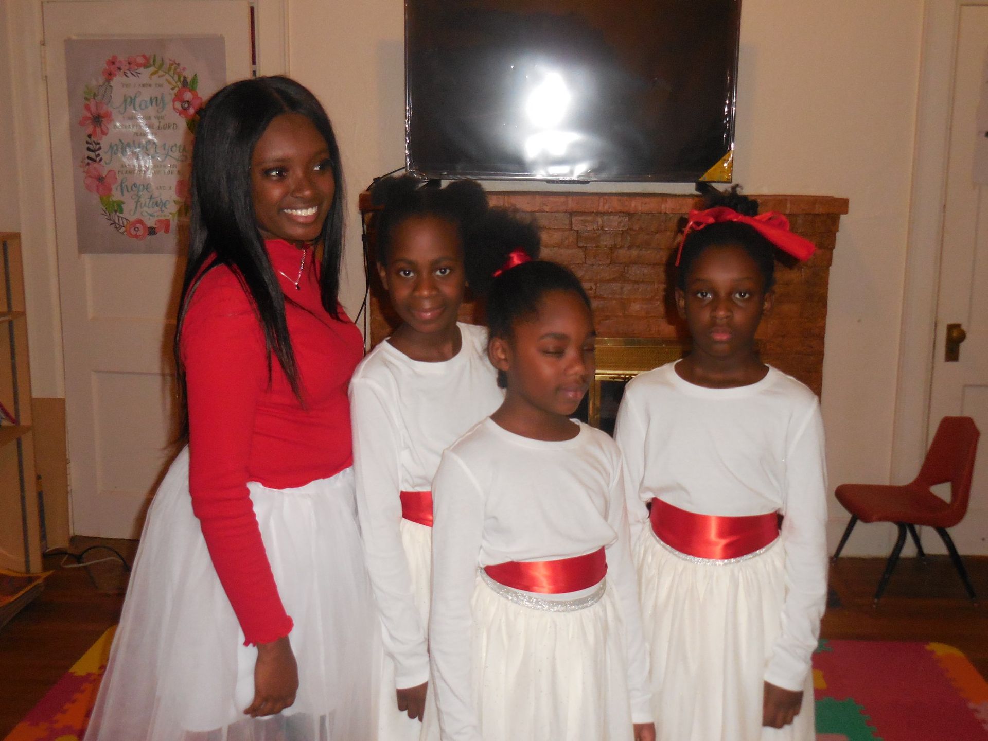 Four people stand in a room in front of a fireplace, wearing white skirts and red tops or sashes, smiling for the camera.