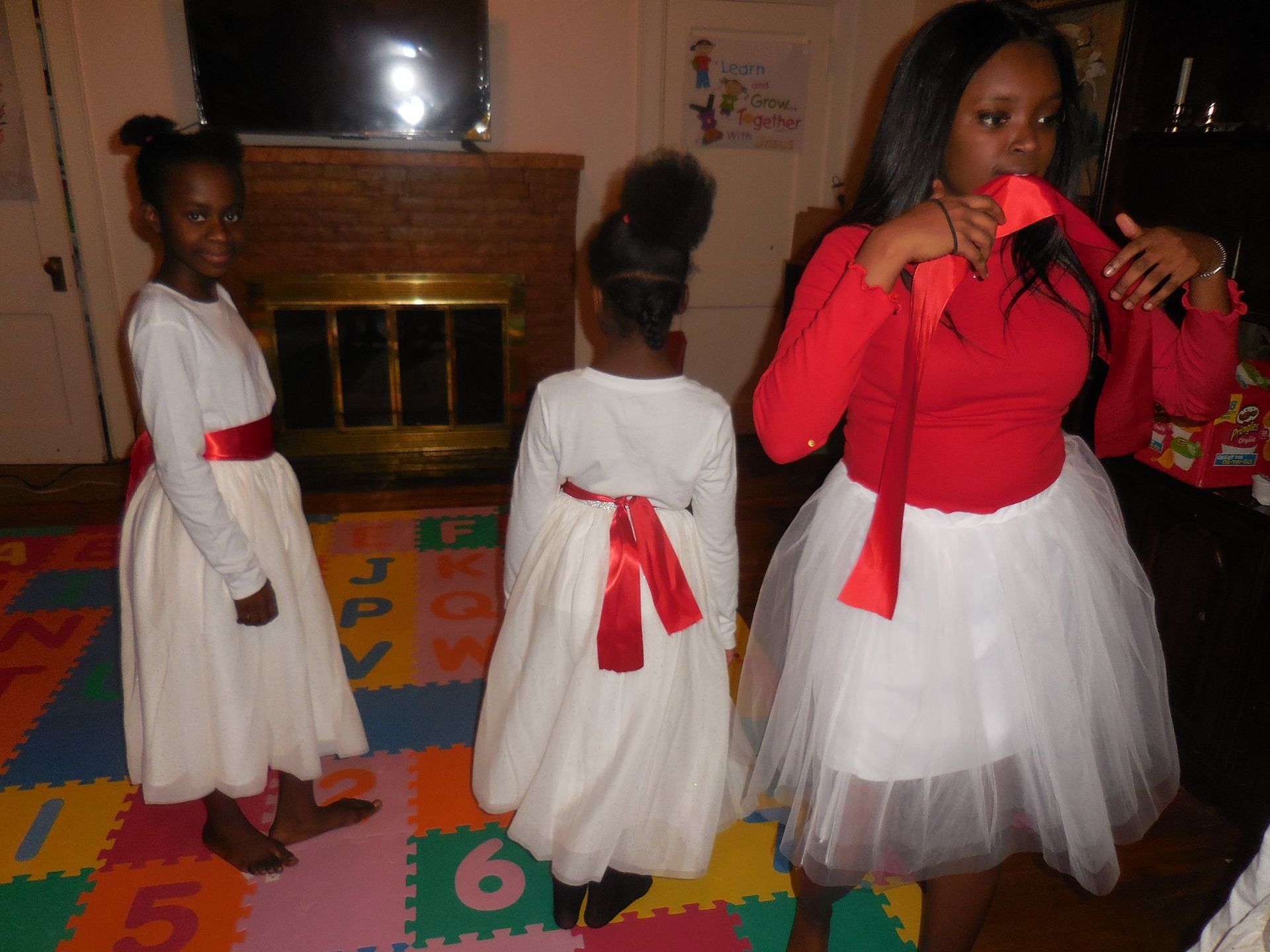 Three people in red and white festive outfits stand in a room with a colorful play mat, fireplace, and television.