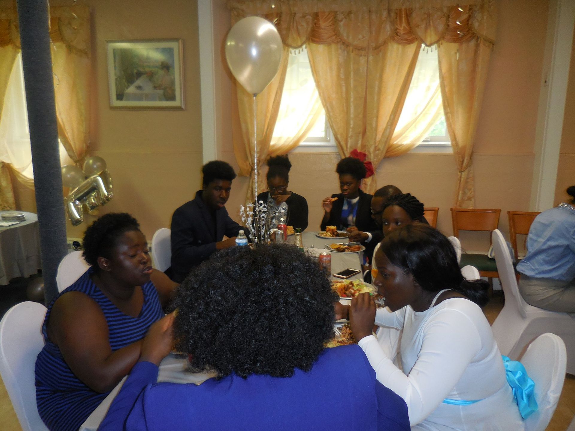 A group of people sits around a round table in an indoor dining area with draped windows, enjoying a meal together.