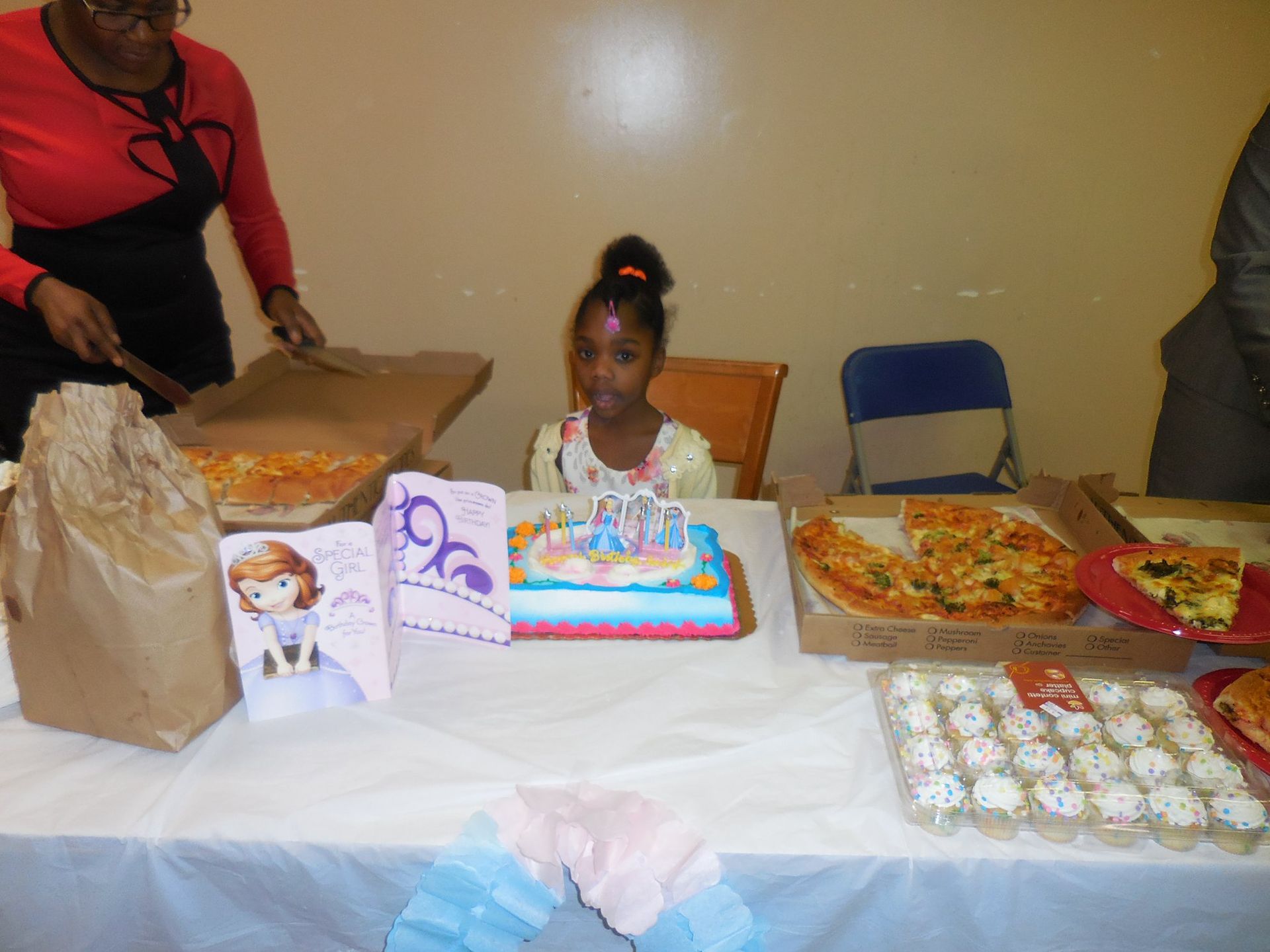 A child sits at a table with a birthday cake, pizza, and party favors, while an adult serves food in the background.