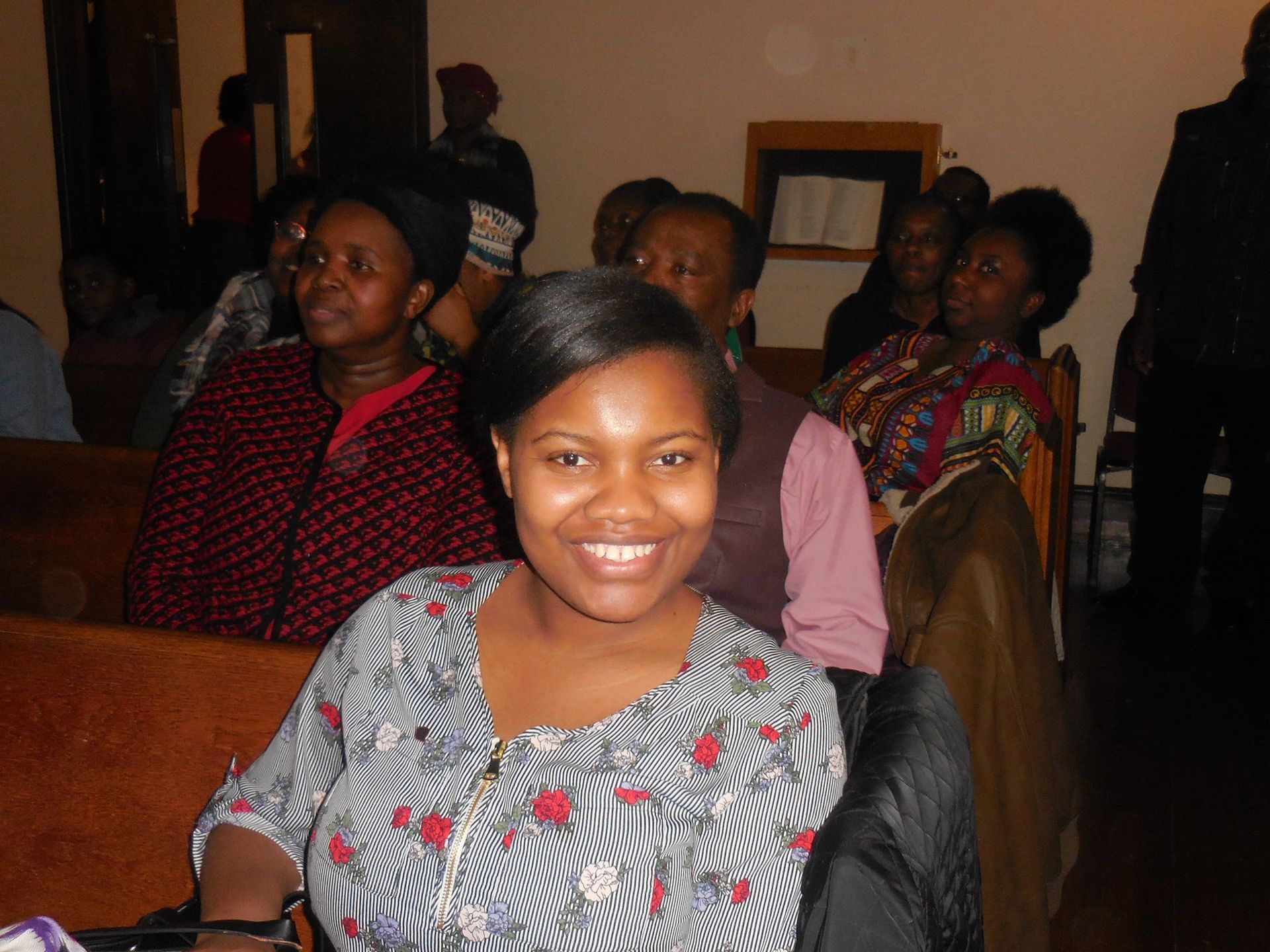 A smiling person in a patterned top sitting in a dimly lit hall filled with seated people.