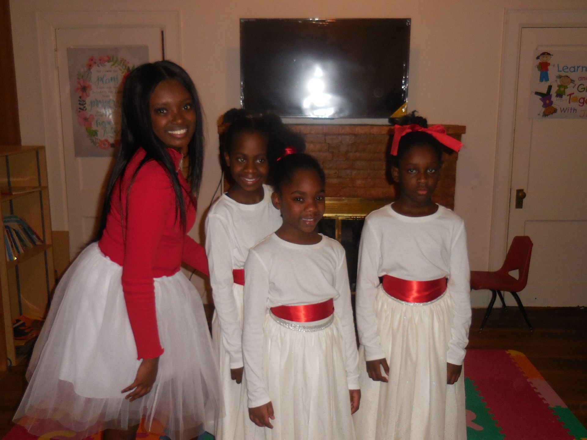 A smiling woman and three children stand together indoors, all wearing white skirts and red tops or sashes.
