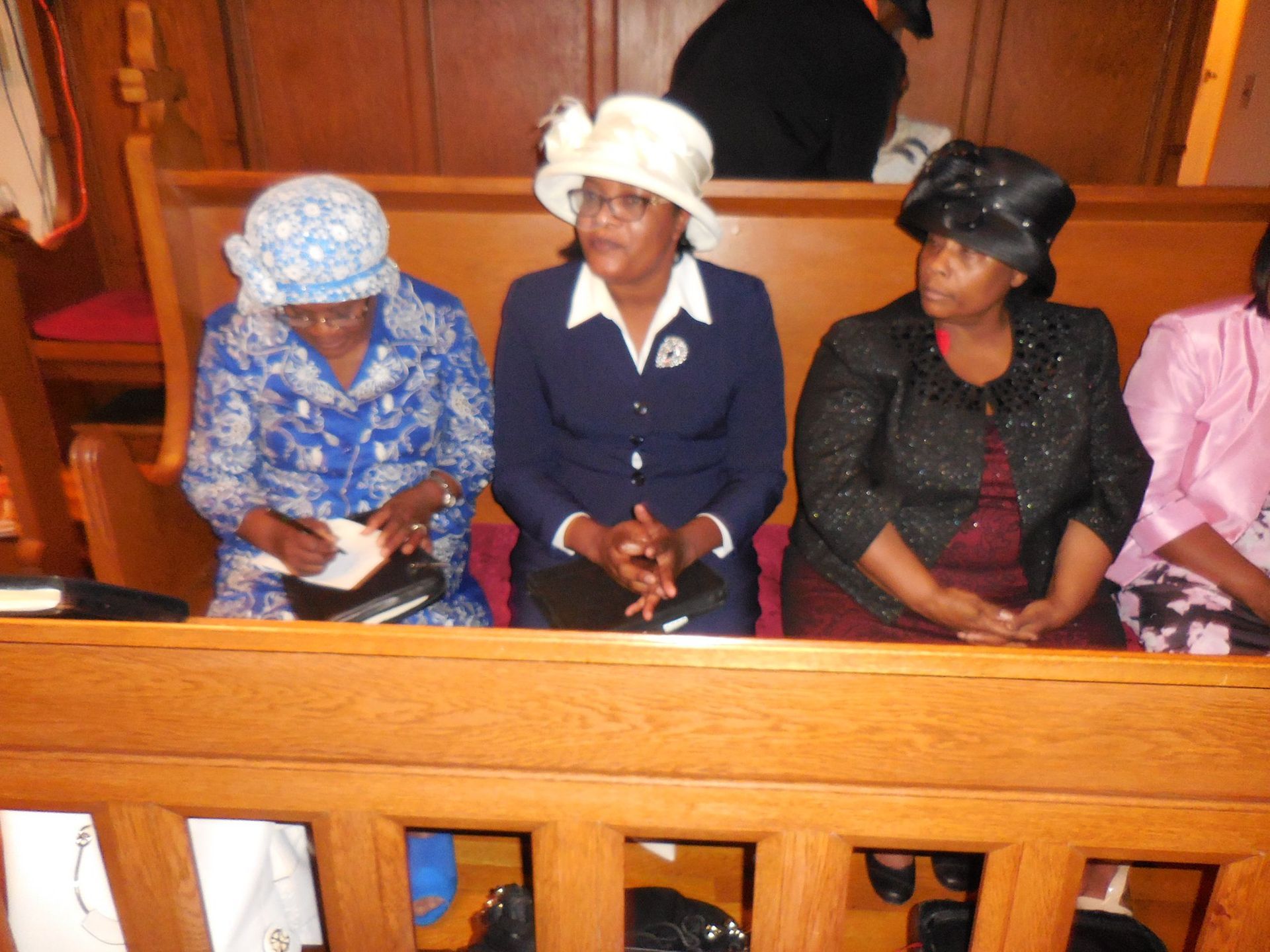 Four women sit in a wooden church pew; one writes in a notebook while the others sit with hands clasped.