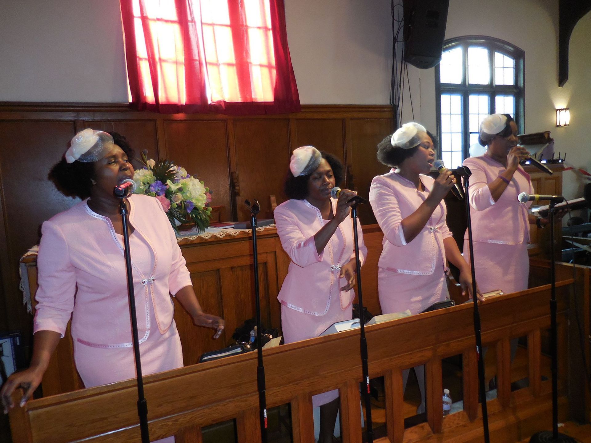 Four singers in matching pink suits and hats perform into microphones in front of a wood-paneled wall and red curtains.