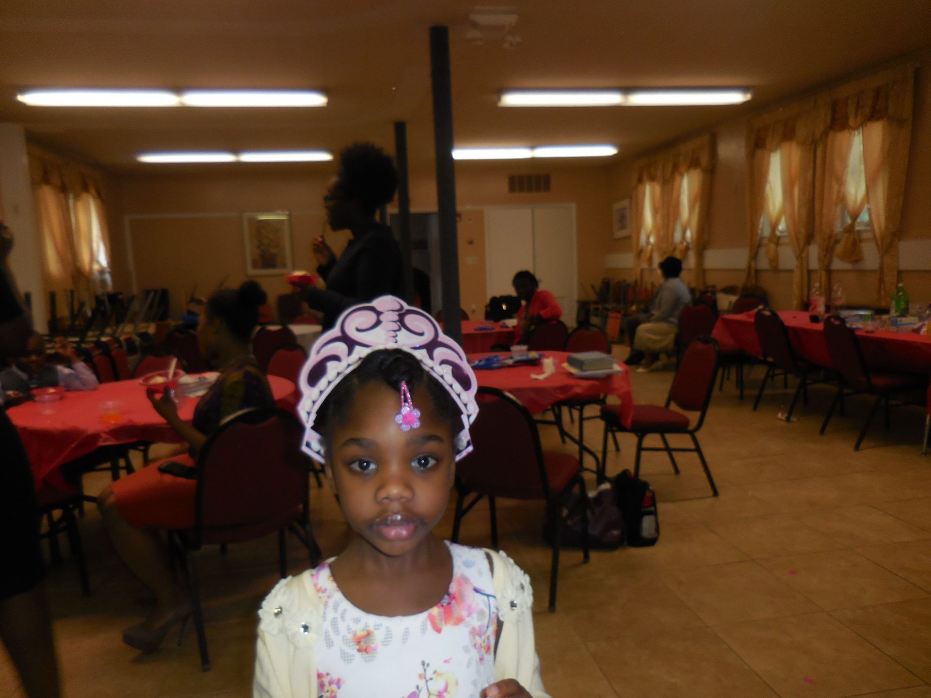 A child wearing a floral top and a decorative headpiece stands in a room with red tablecloths and empty chairs.