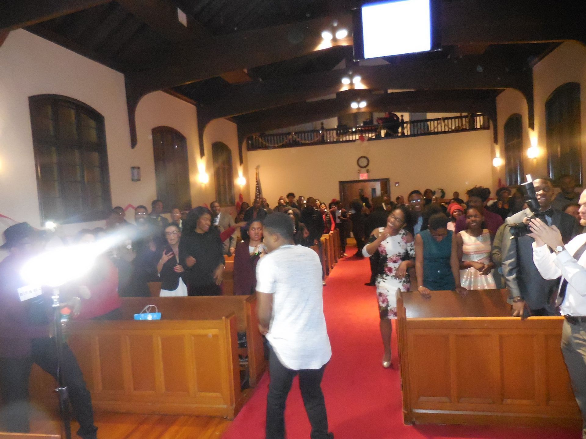A person in a white shirt stands in the aisle of a church, facing a crowd seated in wooden pews during a gathering.