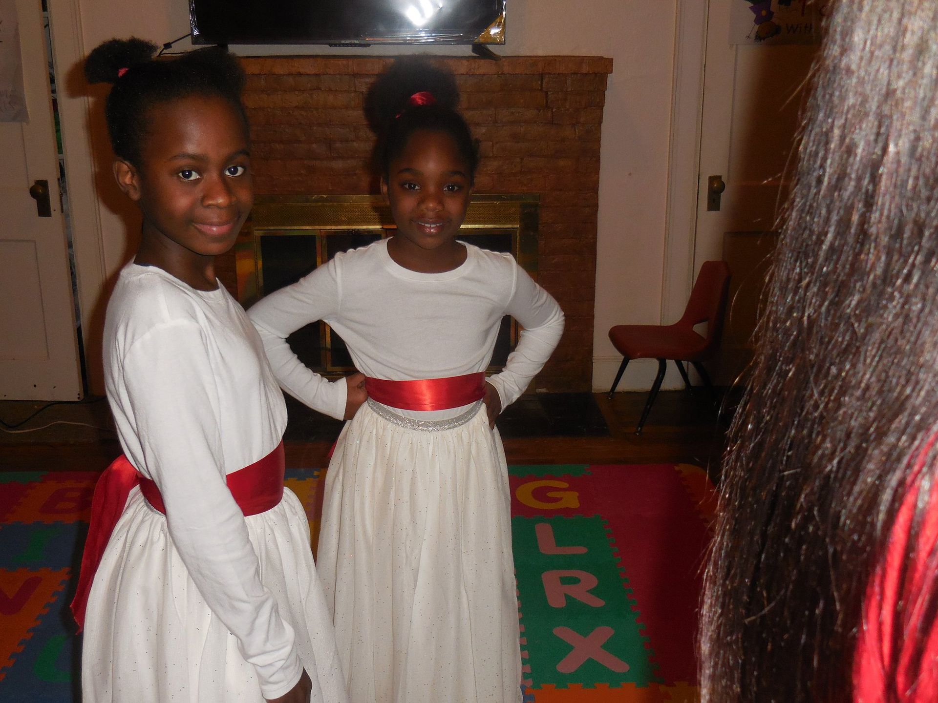 Two children in white long-sleeved dresses with red sashes stand side-by-side in a room with a fireplace and rug.