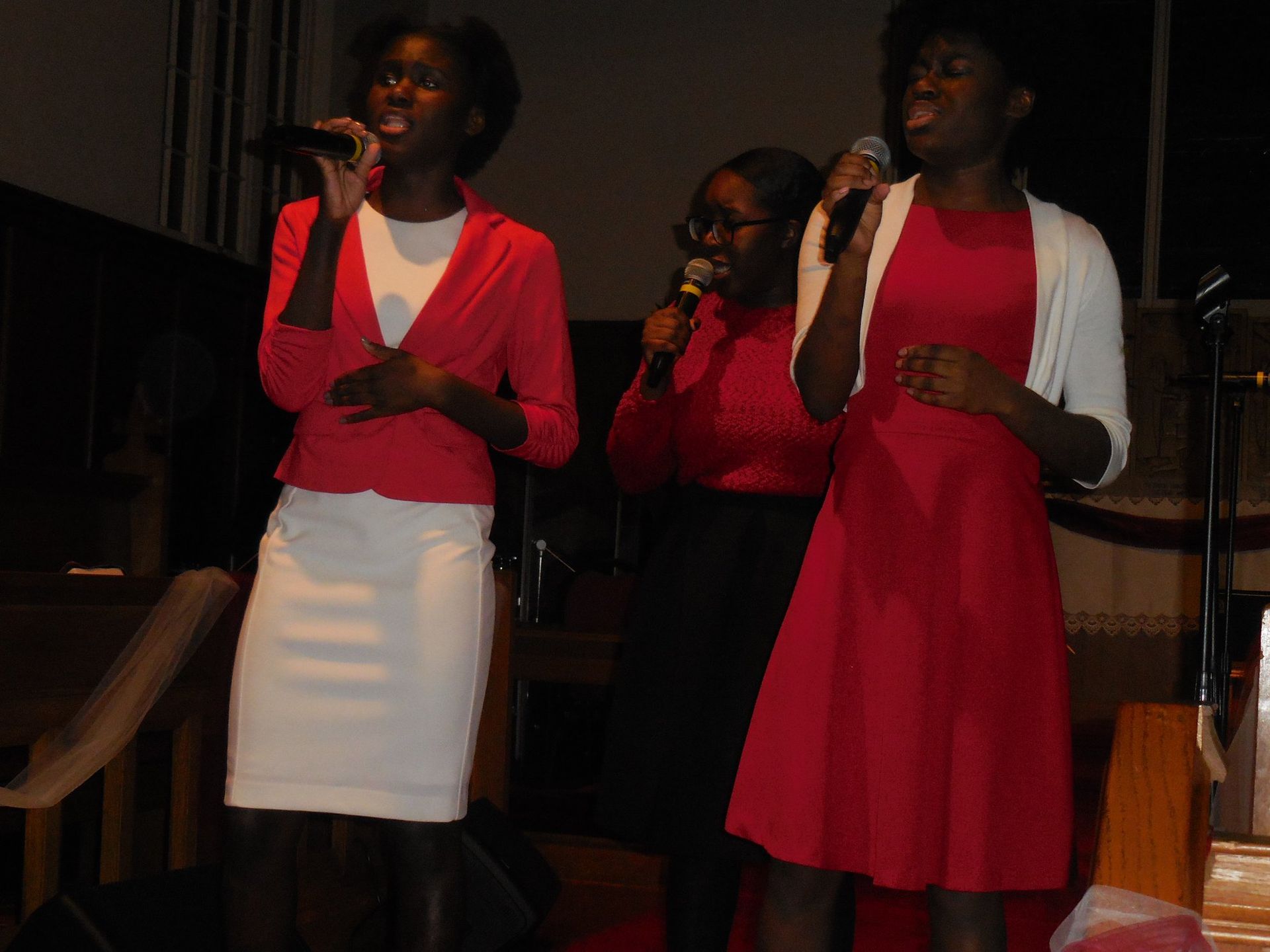 Three people stand on a dimly lit stage, singing into microphones while wearing red clothing.