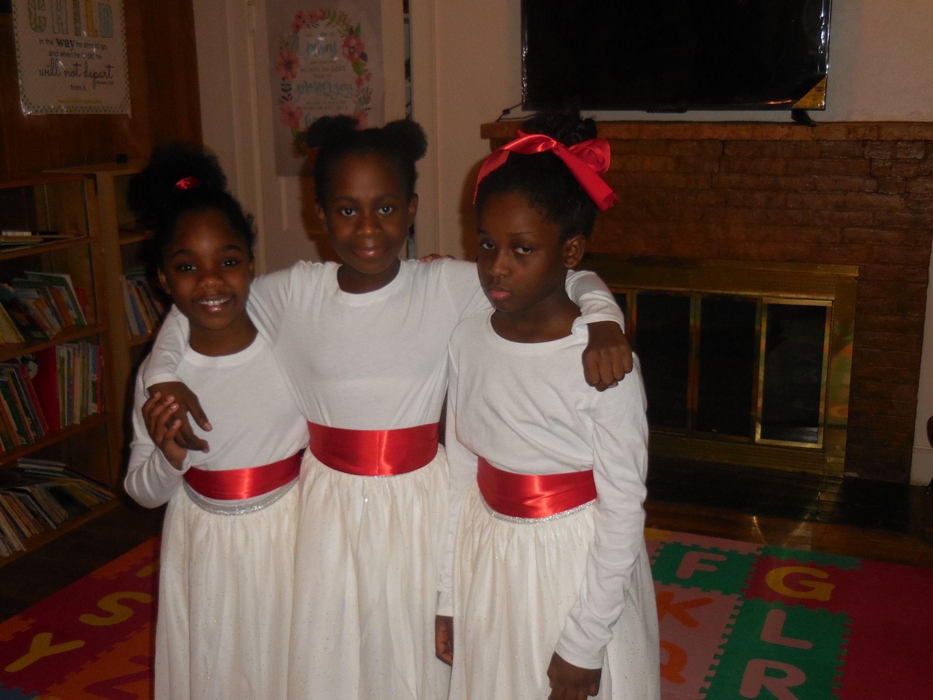 Three people standing together indoors wearing identical white dresses with red sashes, arm in arm near a fireplace.