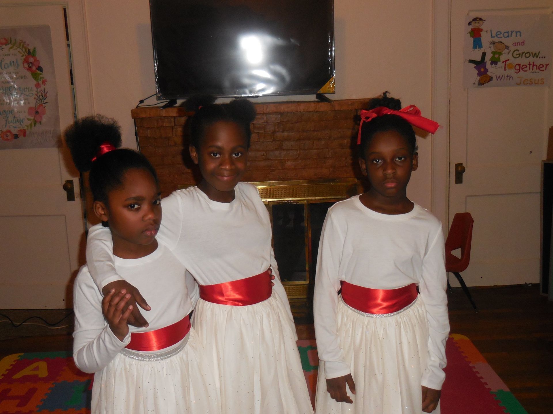 Three children wearing white long-sleeved shirts and white skirts with red sashes stand in a room.