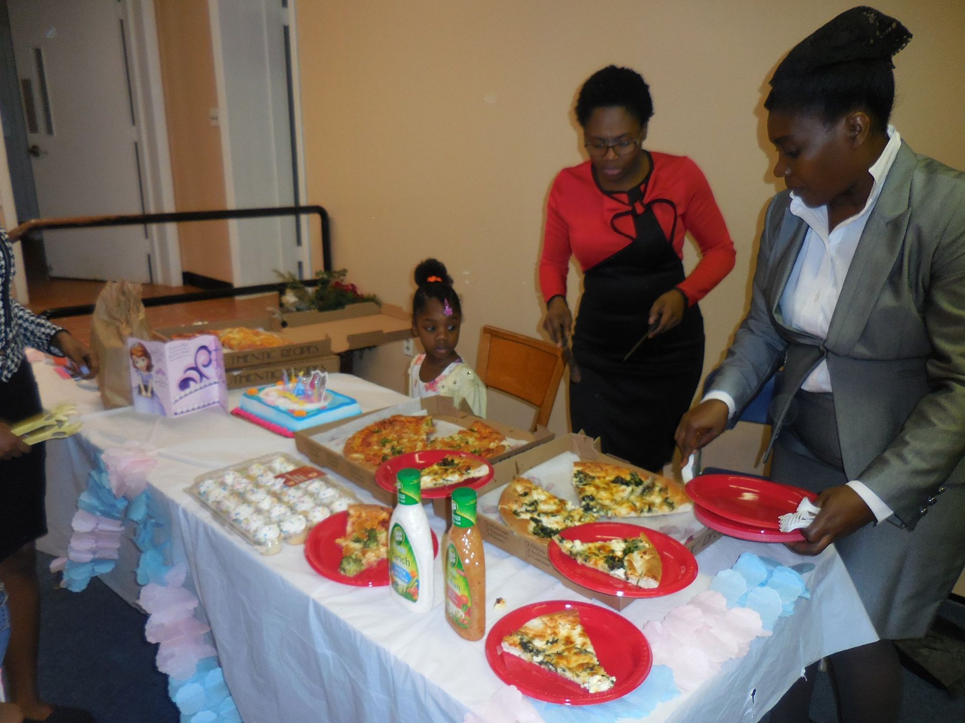 People serving slices of pizza and other party food from a table decorated with pink and blue streamers.