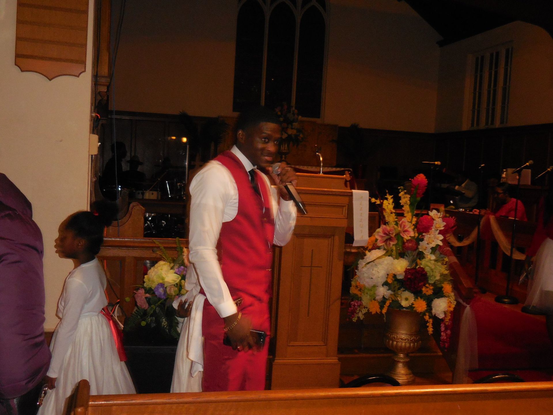 A person in a white shirt and red vest stands at a wooden pulpit, holding a microphone in a church setting.