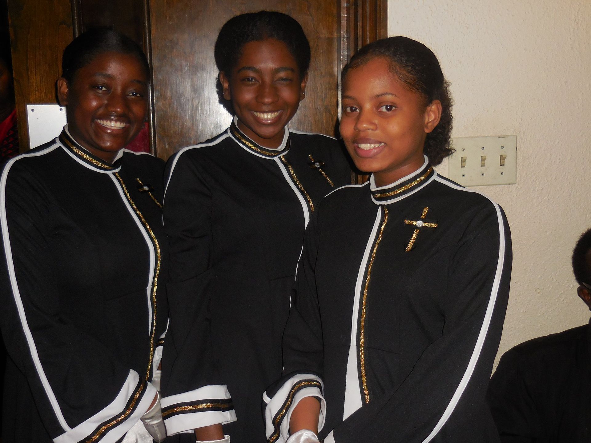 Three people smiling in black choir robes with white trim and gold cross pins, standing indoors.