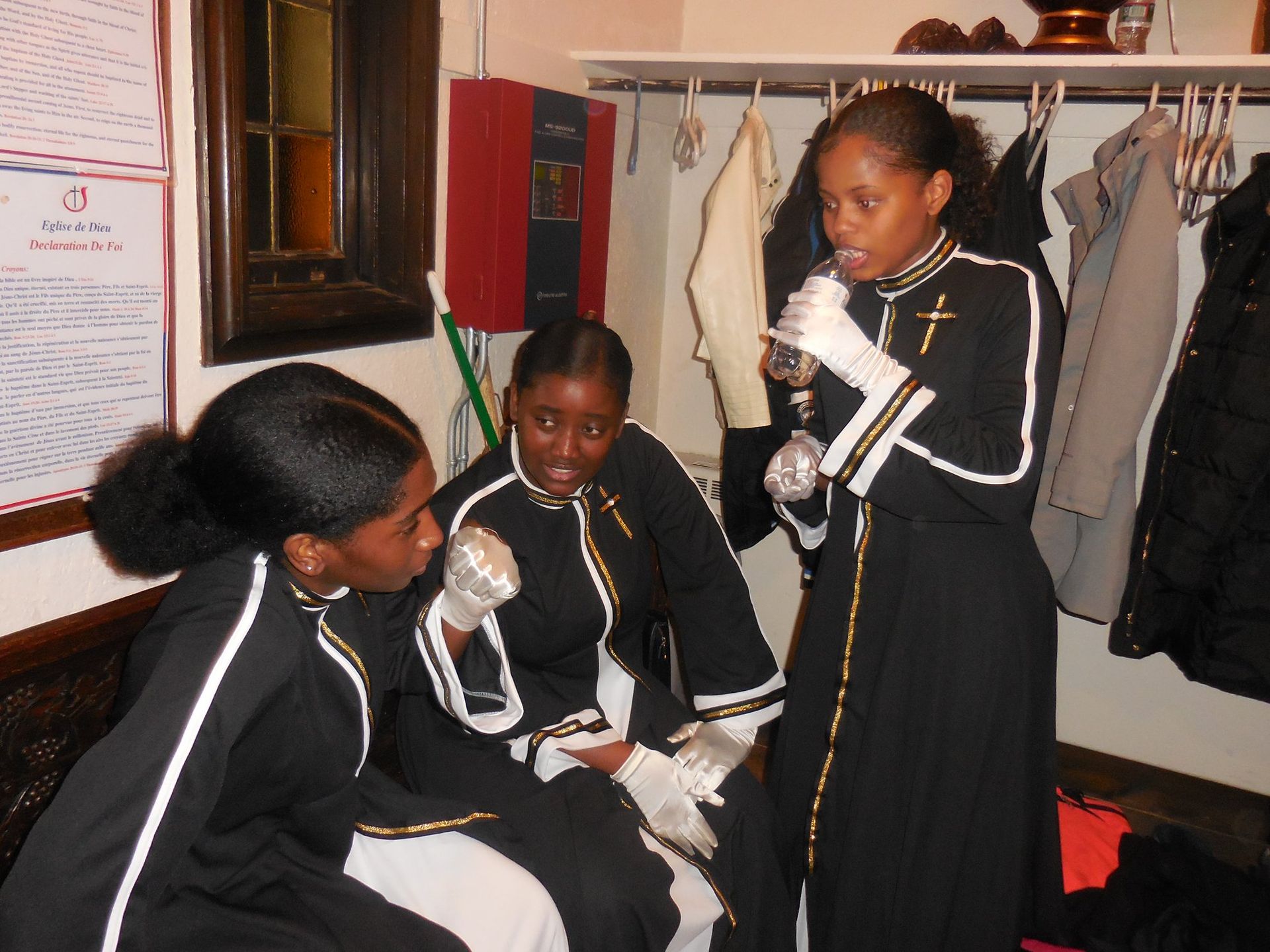 Three people in matching black and white formal choir robes talk in a room with coats hanging on a rack.