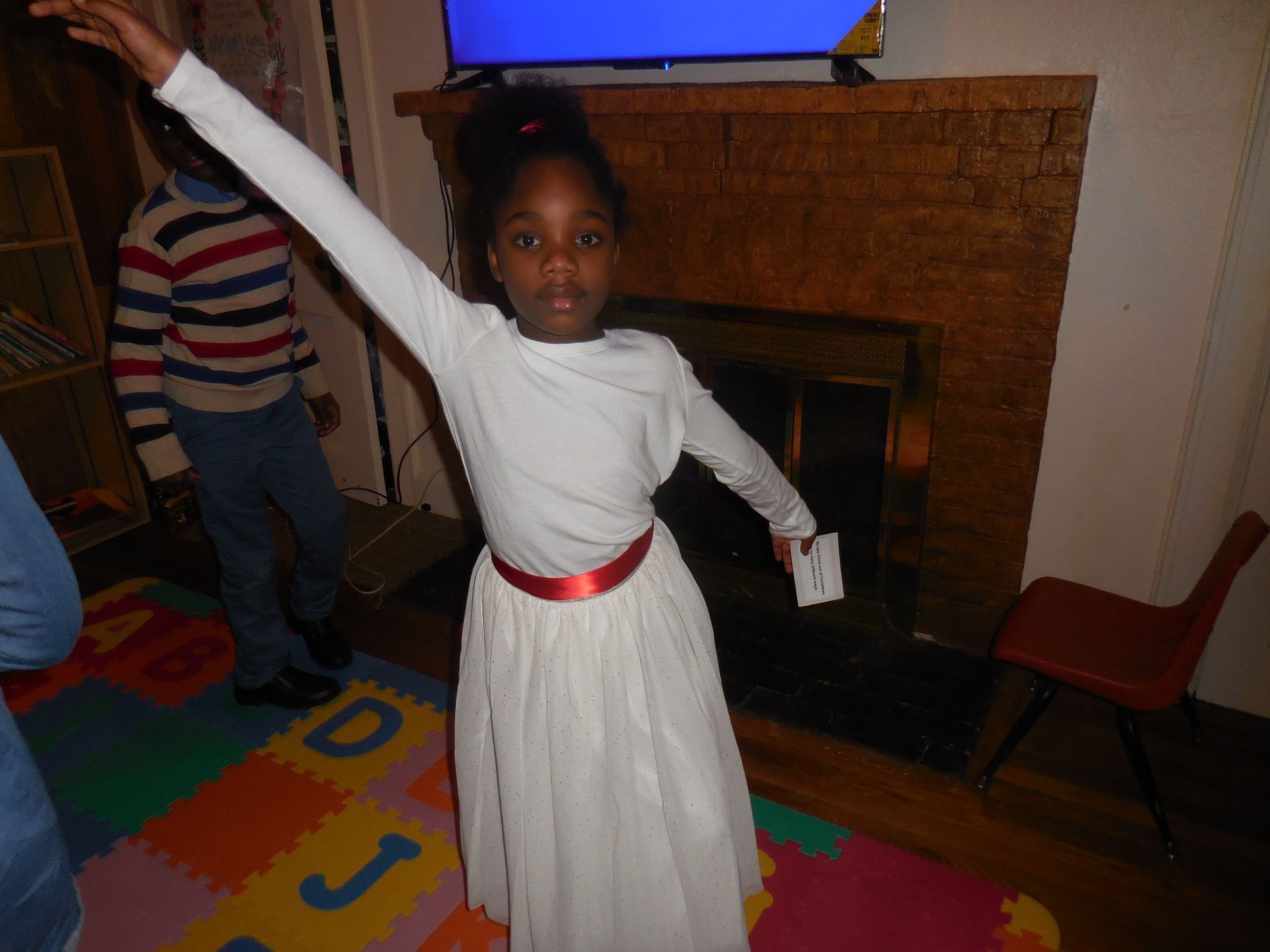 A person in a white long-sleeved shirt and white skirt with a red belt stands with one arm raised in a living room.