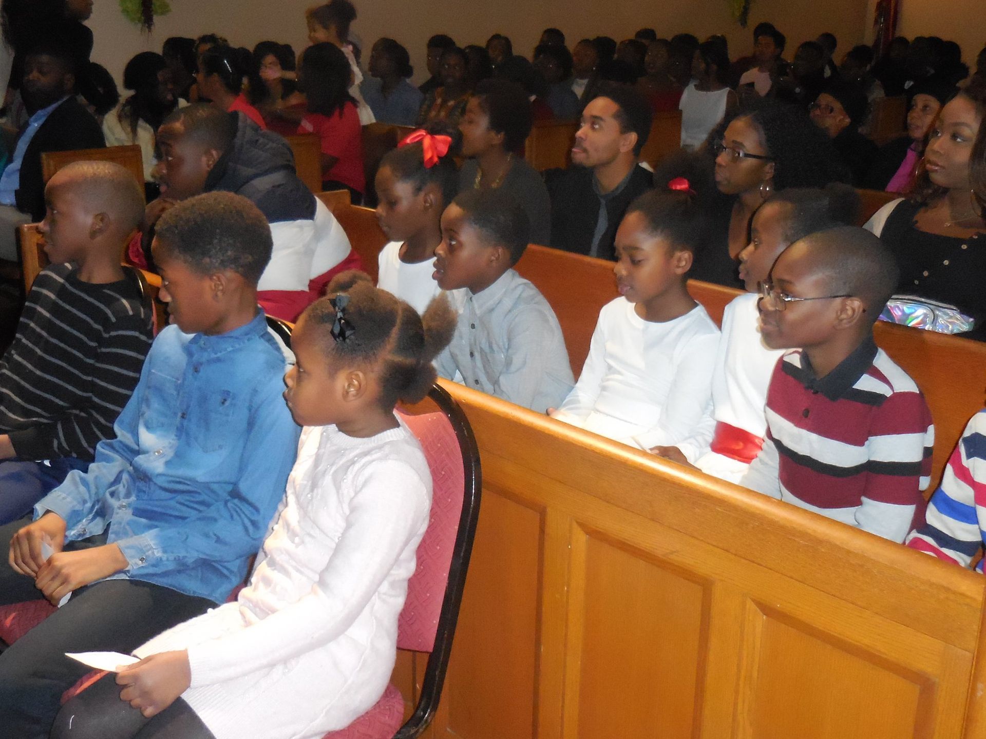 A group of people sitting in wooden church pews, facing forward, dressed in casual clothing.