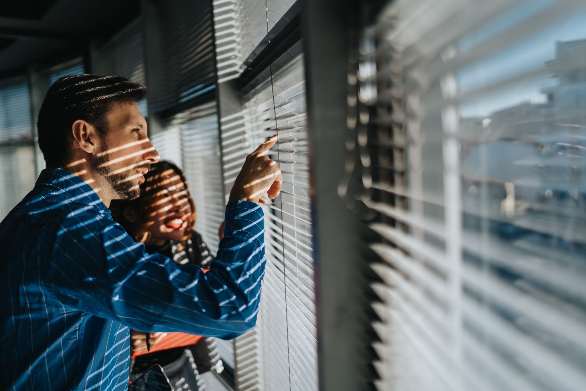 Two people looking through a set of blinds at the city beyond
