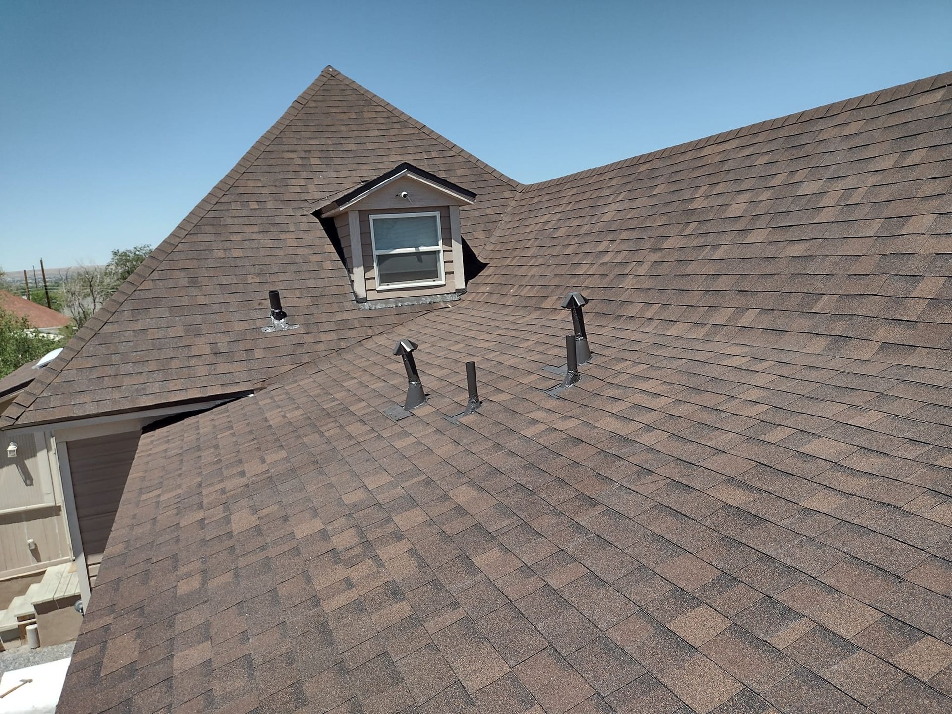 Brown shingled roof of a house with a dormer and vents against a blue sky.