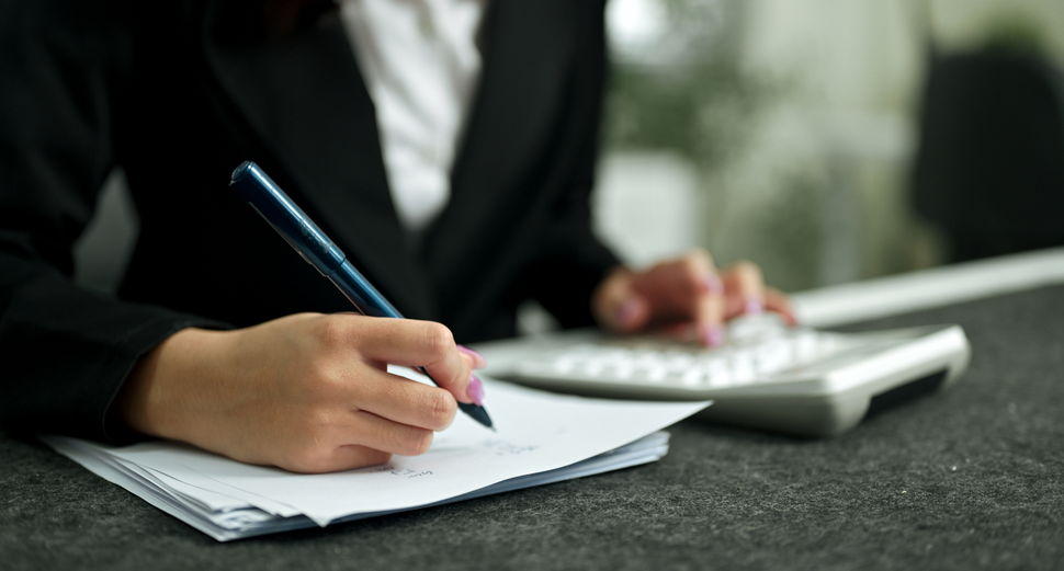 A person in a professional suit writes on paper with a pen while their other hand rests on a calculator.
