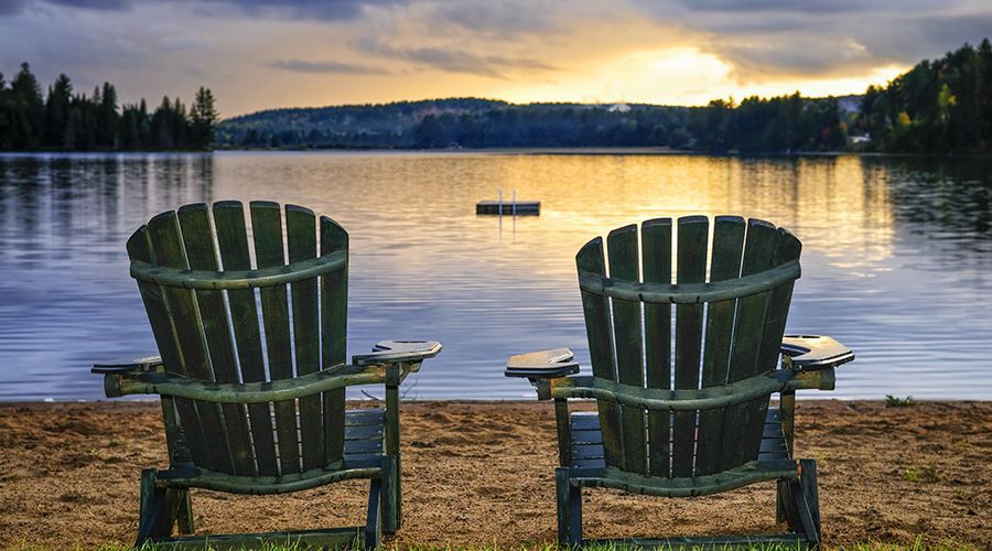 Two wooden chairs are sitting on the beach near a lake at sunset.