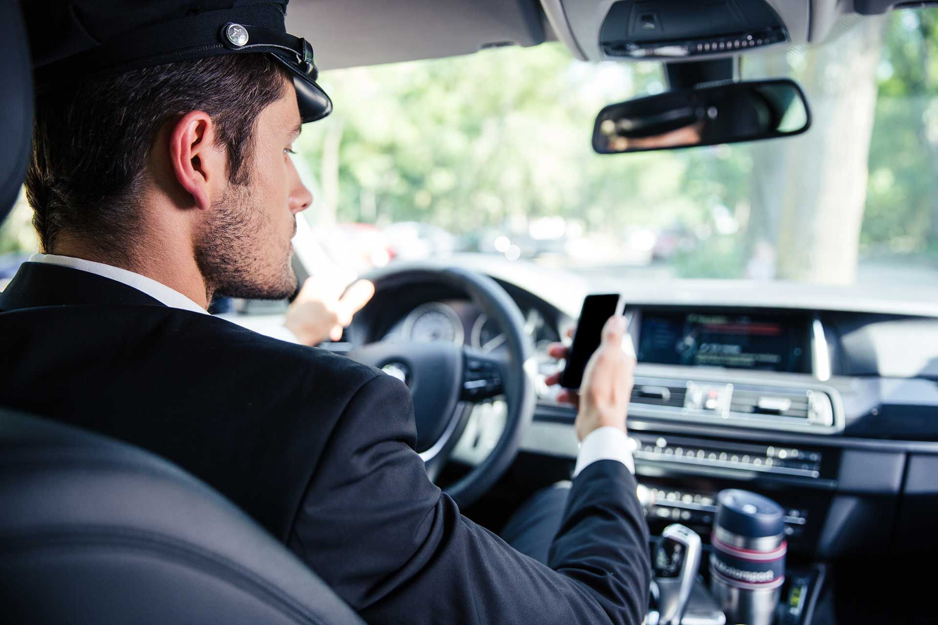 A driver in a suit and cap is sitting in the driver's seat of a car, holding a smartphone while steering.