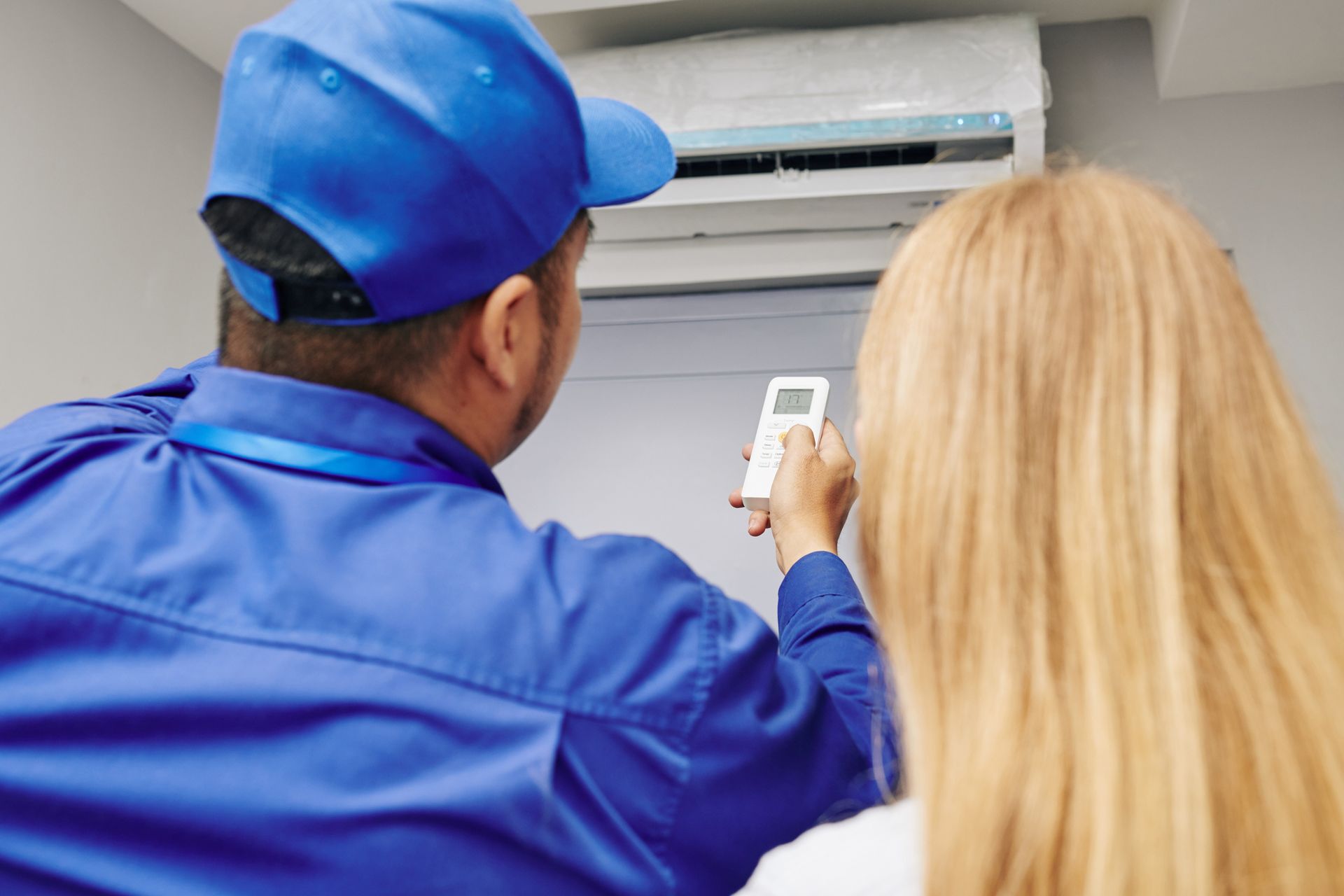 HVAC technician in blue uniform and cap adjusts a wall-mounted air conditioner while a person with blonde hair watches.
