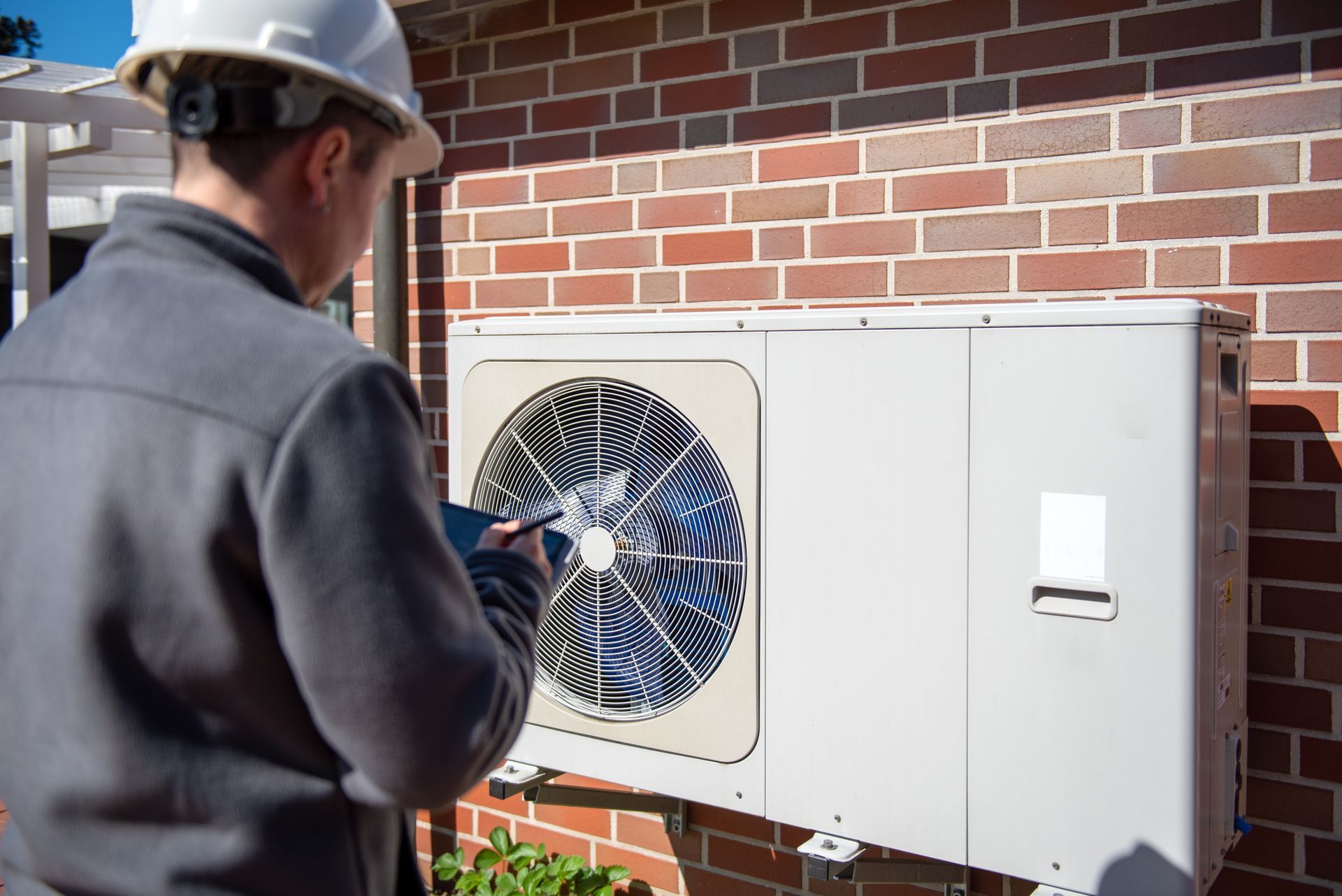 A person in a hard hat inspects an air conditioning unit mounted on a brick wall.