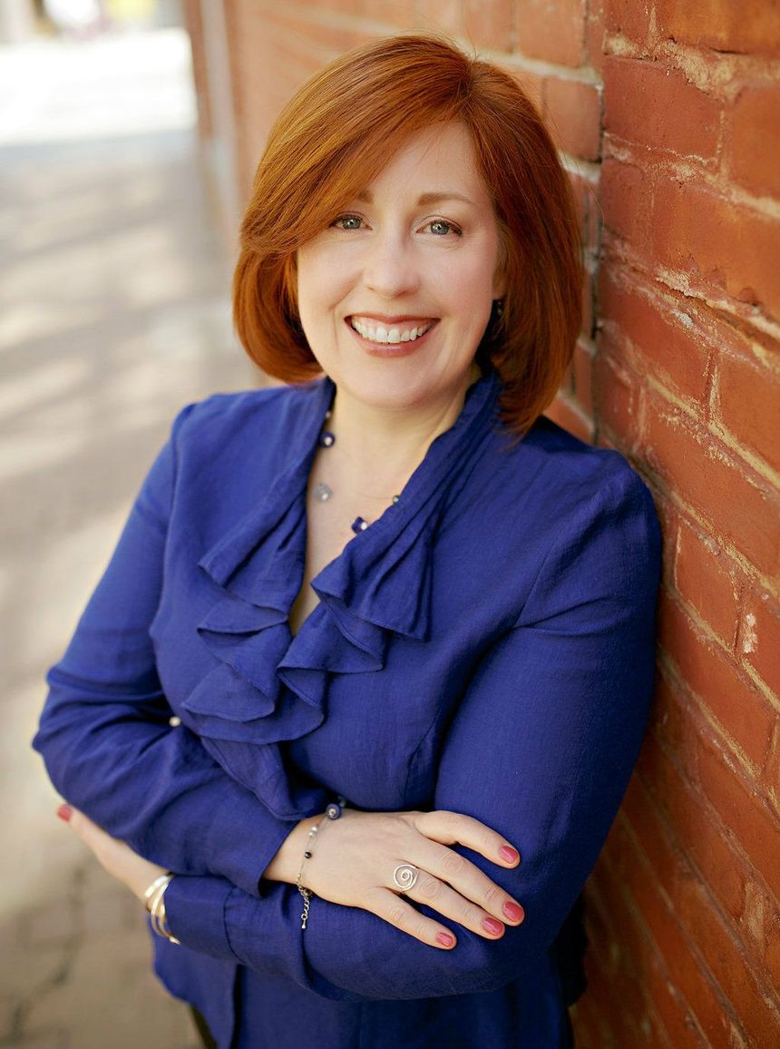 Woman with red hair, smiling in a blue top, arms crossed, leaning against a red brick wall.