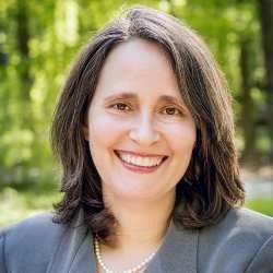 Woman with brown hair smiles, wearing a pearl necklace and gray blazer, outdoors.