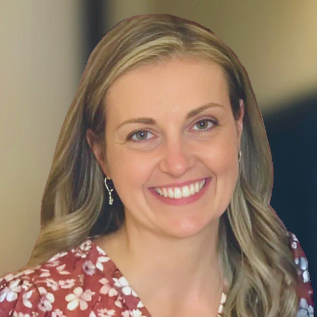 Blond woman smiles at camera, wearing a blue shirt, pearl earrings, with a bridge and water in the background.